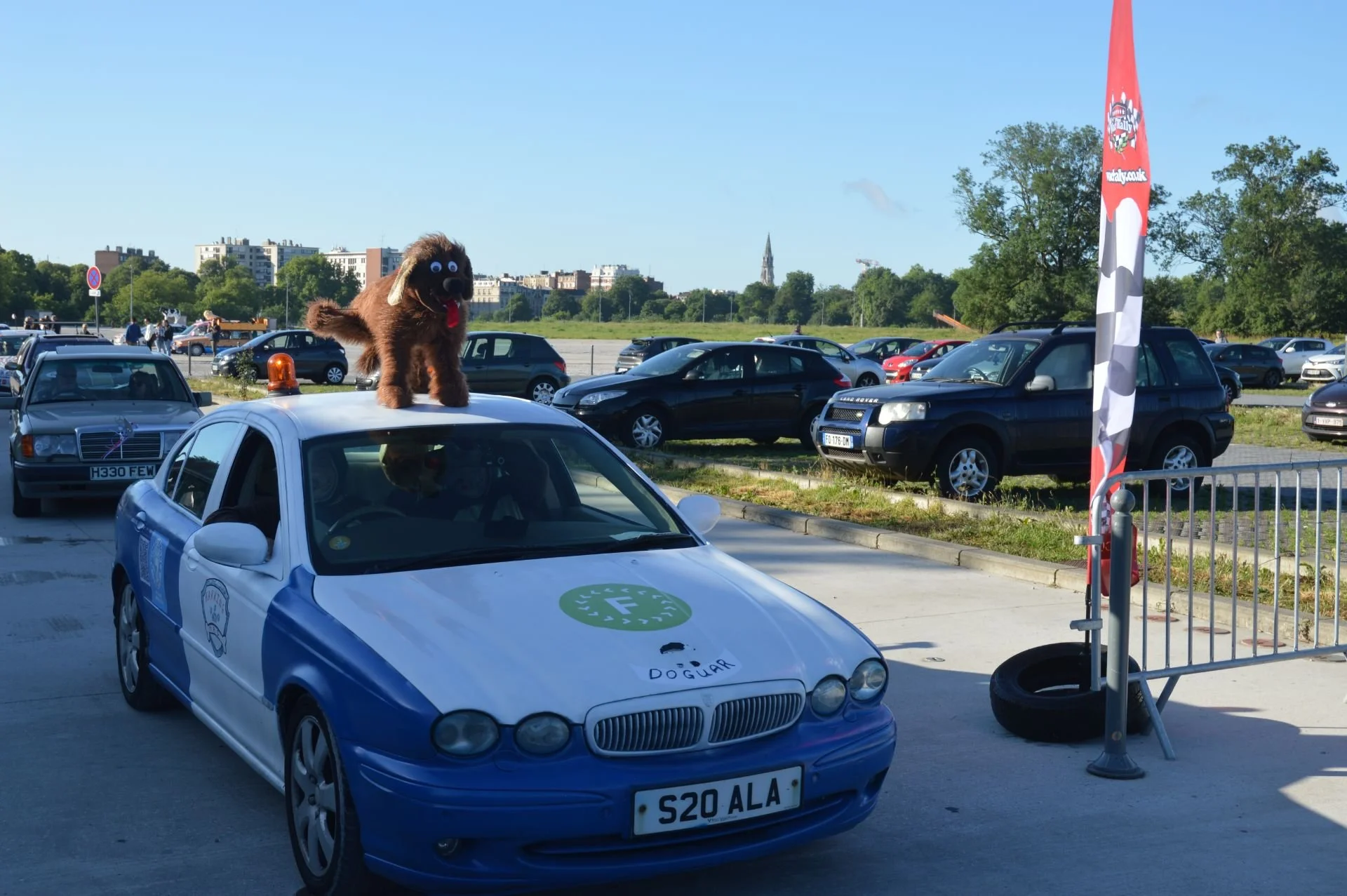 A costumed dog mascot standing on top of a police car in an outdoor parking lot with other parked cars and trees in the background.