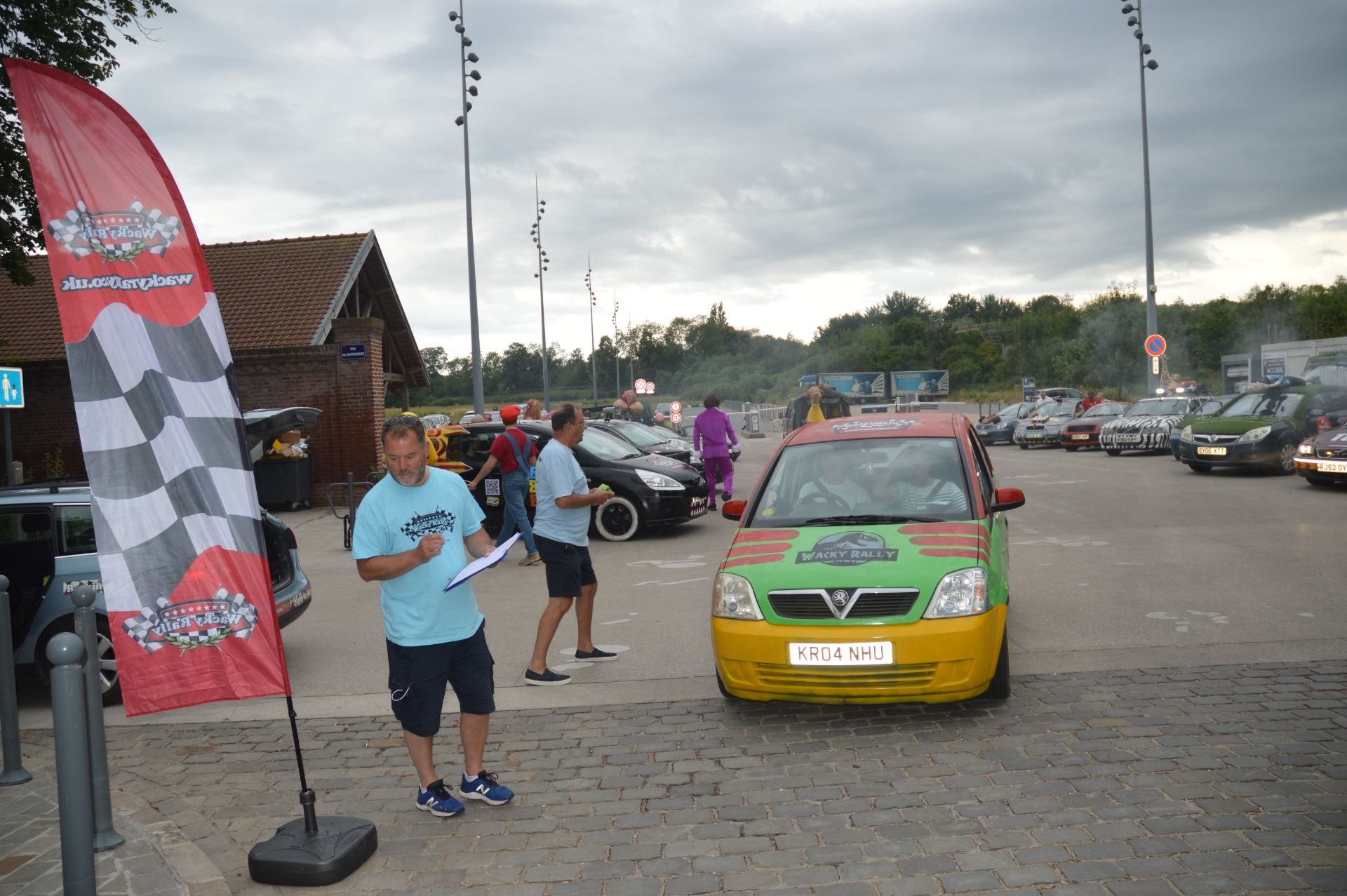 A parking lot at a rally event with modified cars and people walking around, one person reading notes, and a red and green car in the foreground.