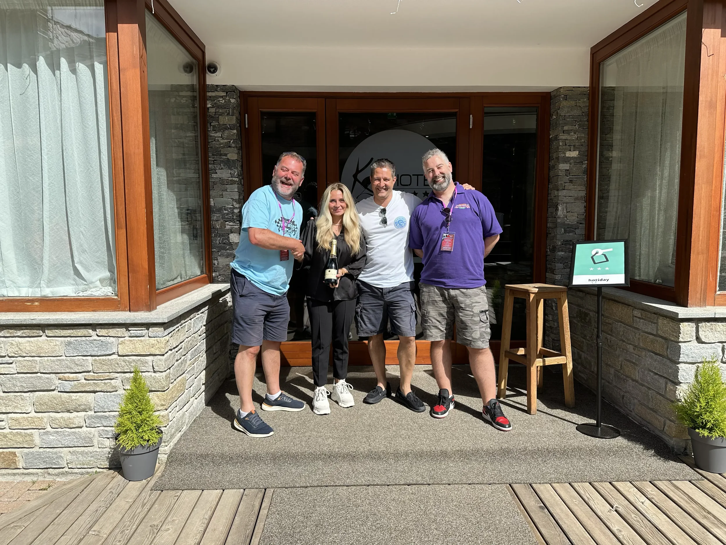 Four people standing outside a hotel entrance smiling, one holding a bottle of champagne, with a small potted plant on each side and a sign indicating 'hotel' with luggage icon.