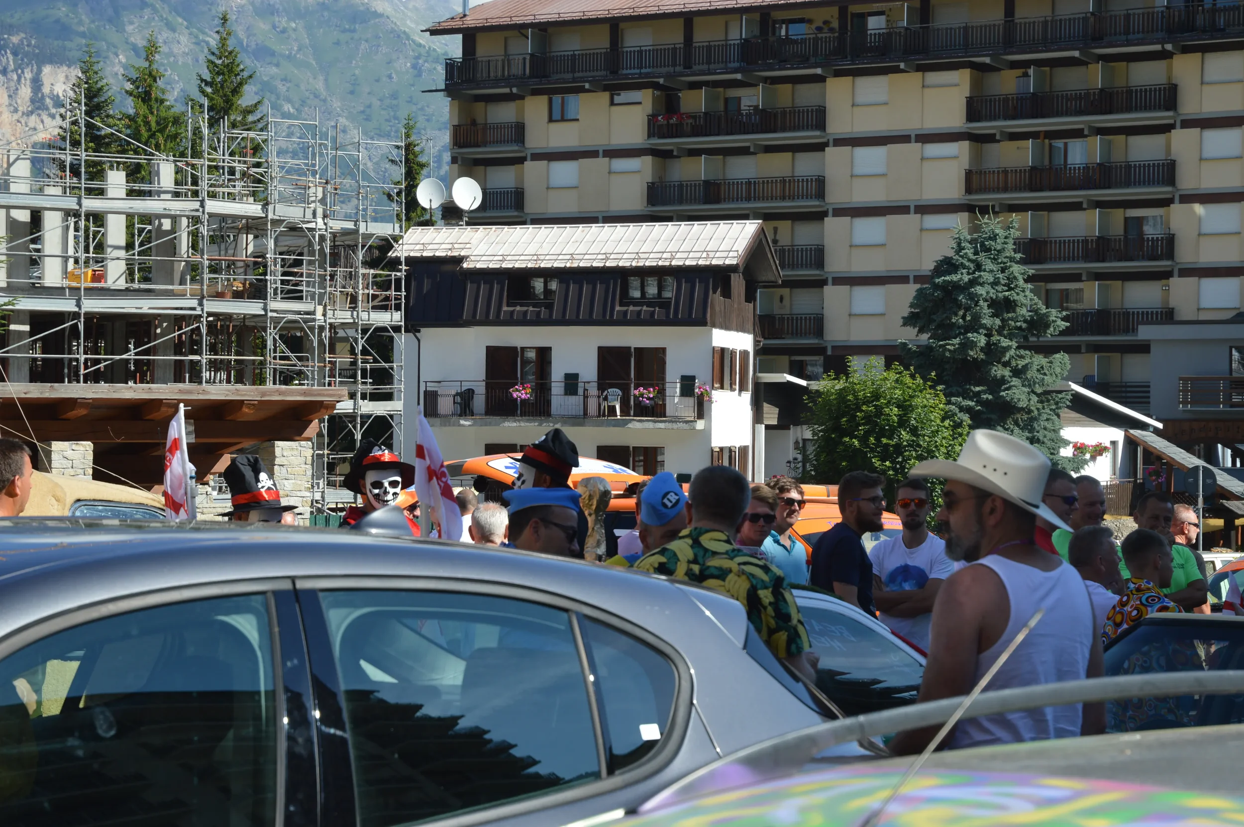 A crowd of people gathered outdoors, with some dressed in costumes, in front of buildings and a mountain backdrop, at what appears to be a festival or parade.