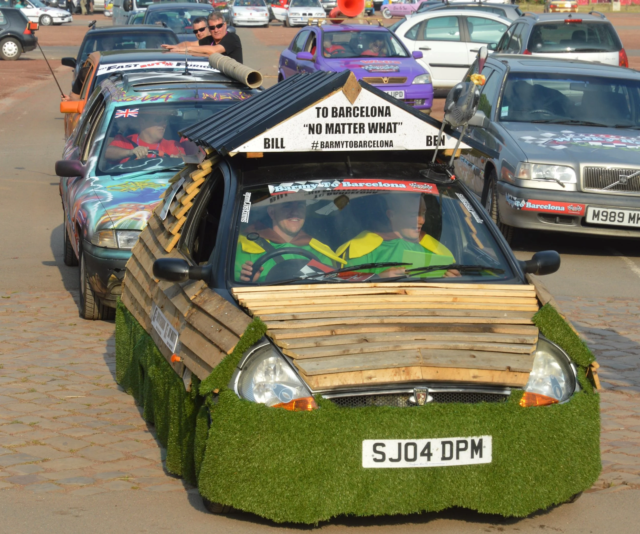 A small car decorated with wooden planks on the hood, artificial grass on the sides, and a sign on the roof that reads 'To Barcelona 'No Matter What' Bill.' The car has people inside wearing green jackets, and it is part of a parade or event involving decorated cars, with other vehicles visible in the background.