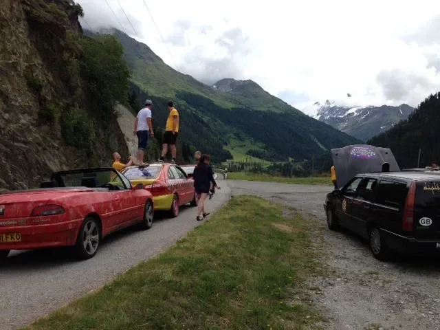 Multiple cars parked on a gravel roadside with people sitting and walking on and around the cars, surrounded by green mountains and cloudy sky.
