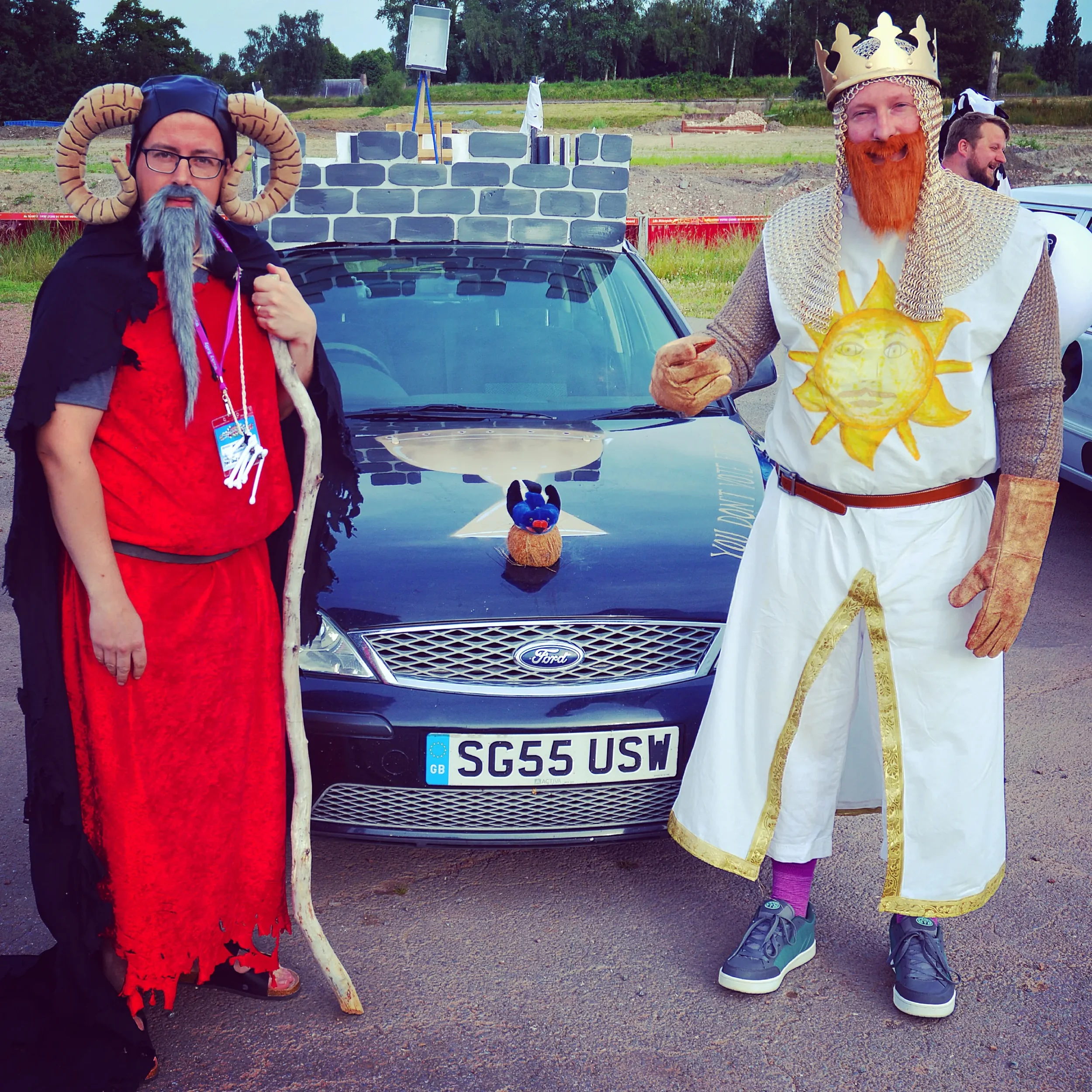Two people dressed as mythical or fairy tale characters standing in front of a black Ford car with a small plush toy on the hood. One is dressed as a wizard or monster with a horned headpiece, glasses, gray beard, and red and black costume. The other