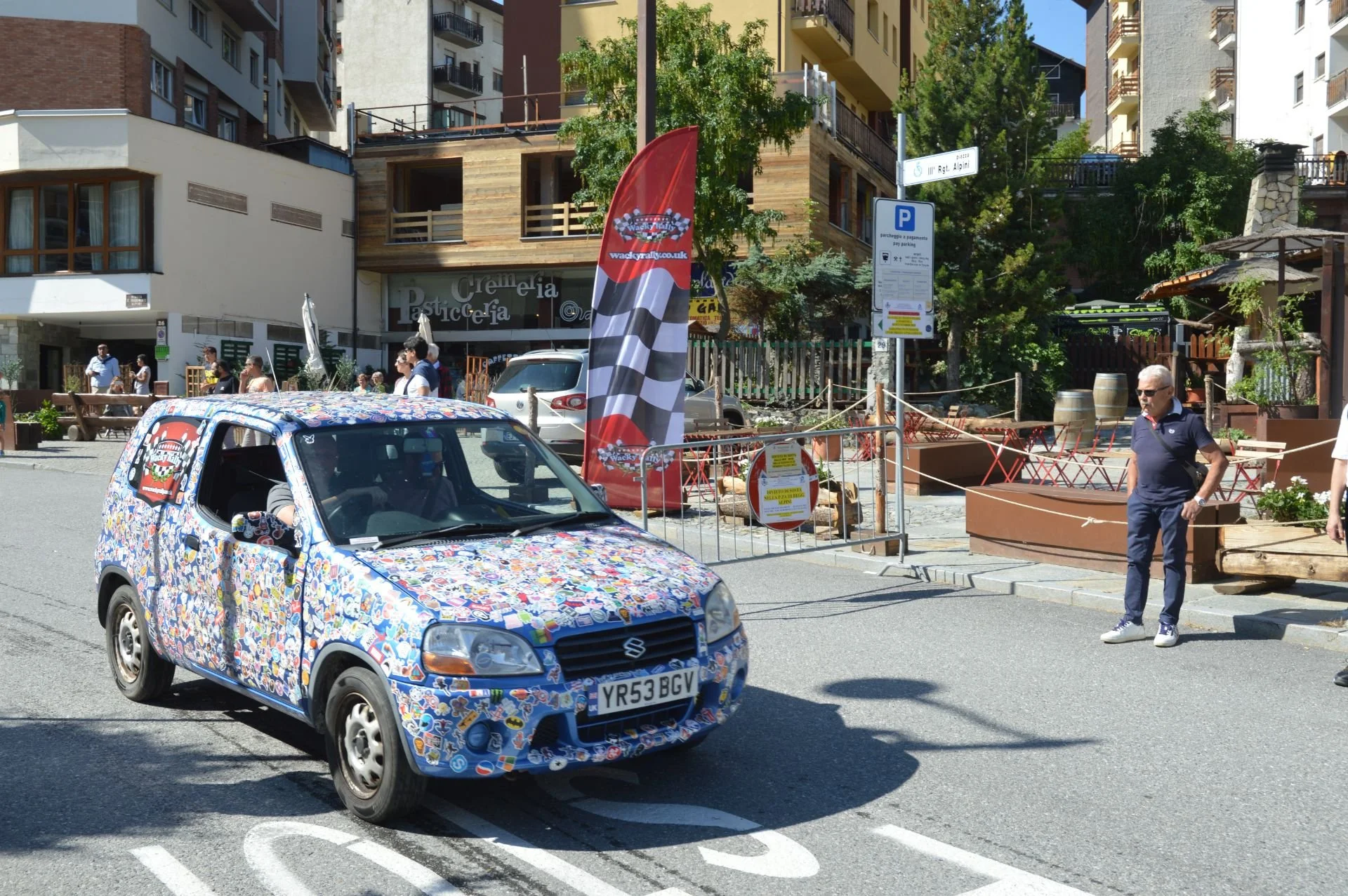 A street scene with a small car covered in colorful stickers parked near a sidewalk with a man standing nearby. There are buildings with outdoor seating in the background, and a red and black flag with a checkered pattern and the word "Wacky" on it, 