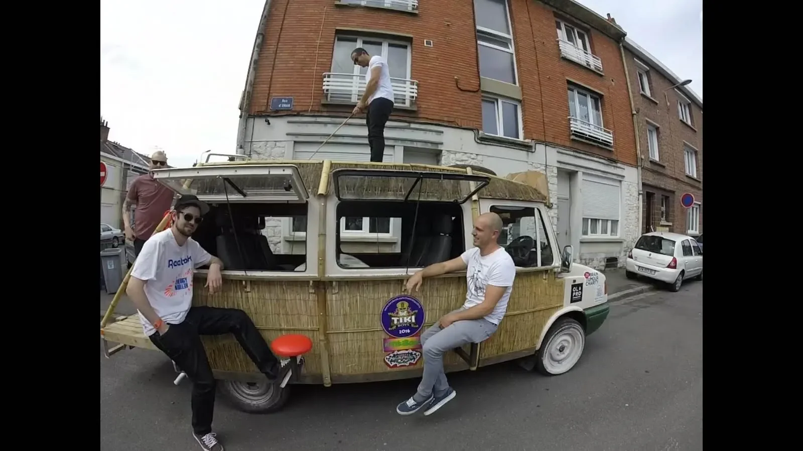 Four men with a vintage van decorated like a tiki hut on a city street. One man stands on top of the van cleaning the roof, another leans against the side of the van, and two more sit nearby. The background features brick residential buildings.