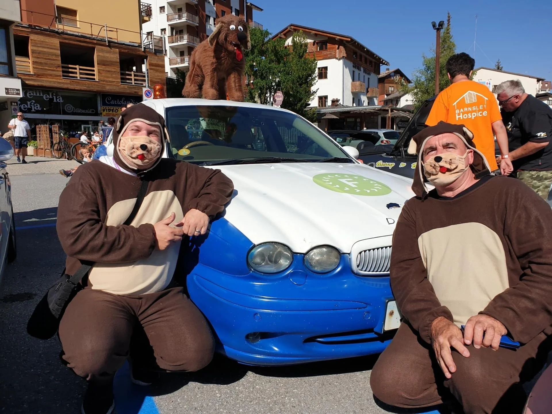 Two people dressed in dog costumes posing in front of a car with a brown dog mascot on the roof. There are other people and buildings in the background, suggesting an outdoor event.