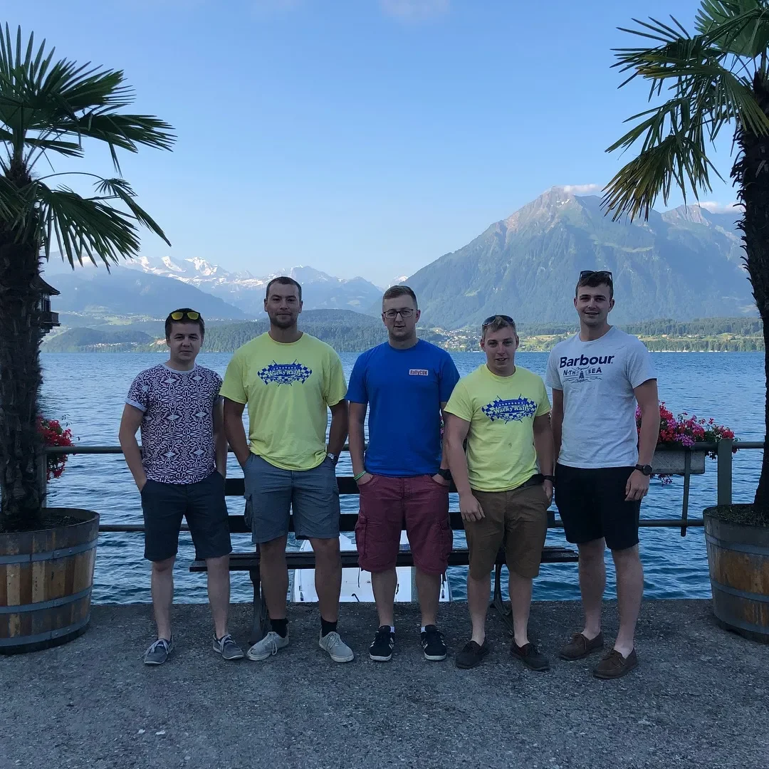 Group of five young men standing outdoors near a lake with mountains in the background, palm trees on either side, and a clear blue sky.