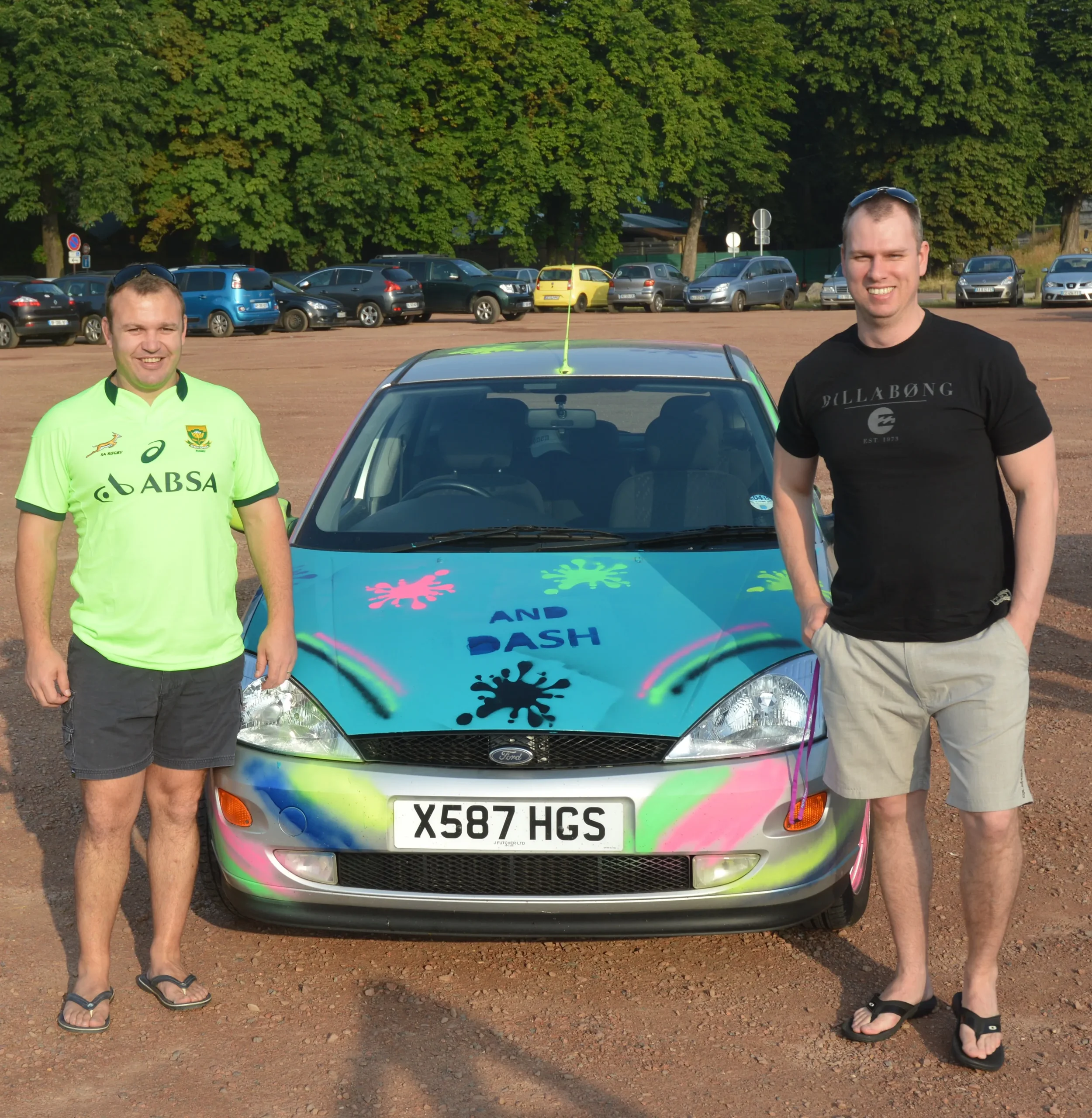 Two men stand next to a colorful car with spray paint designs and the words "AND DASH" on the hood, in a parking lot with other cars and green trees in the background.