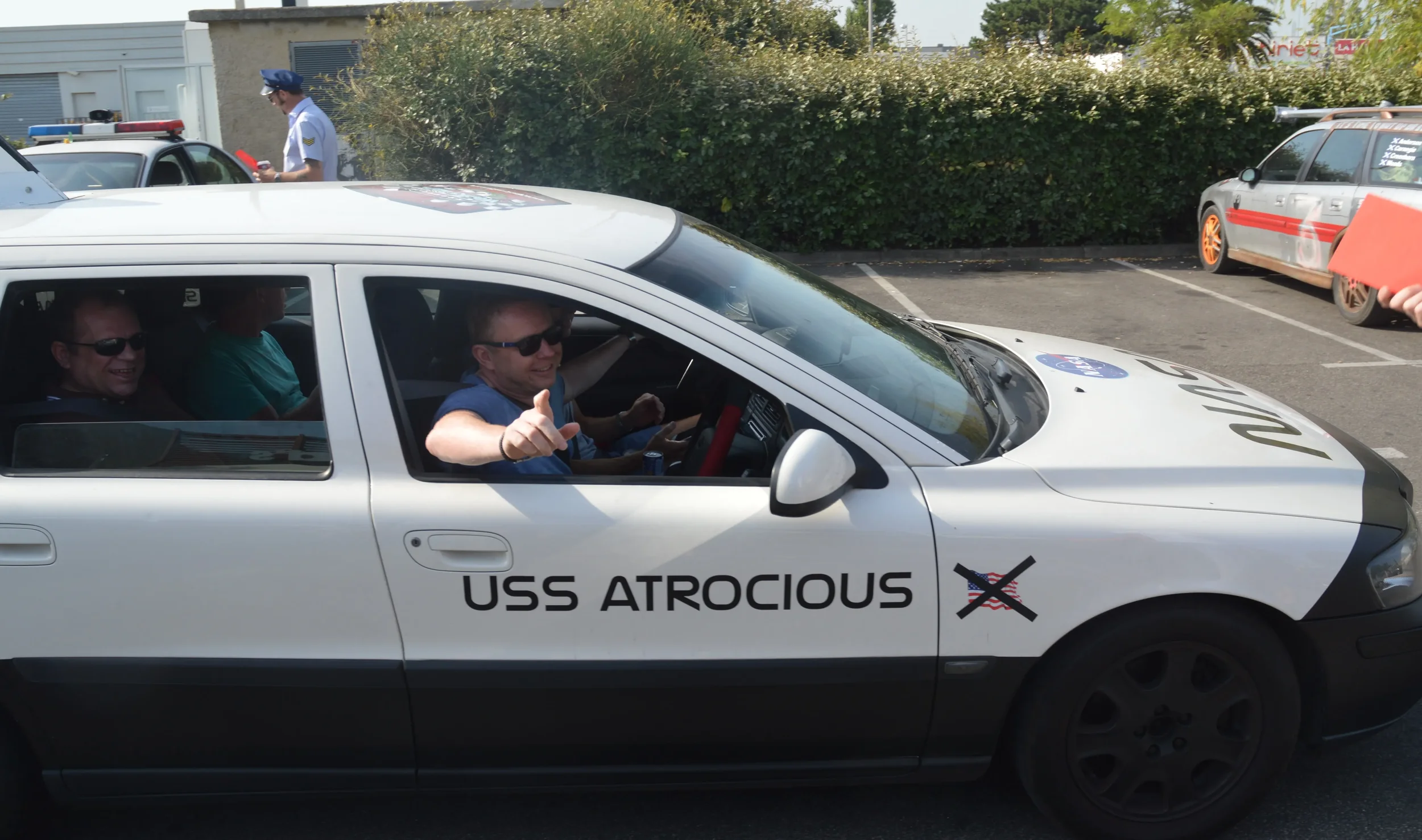 A white car with the text 'USS ATROCIOUS' on the side and a crossed-out American flag. Inside, three people are smiling, with the driver giving a thumbs-up. In the background, there is a person in a uniform and another vehicle.
