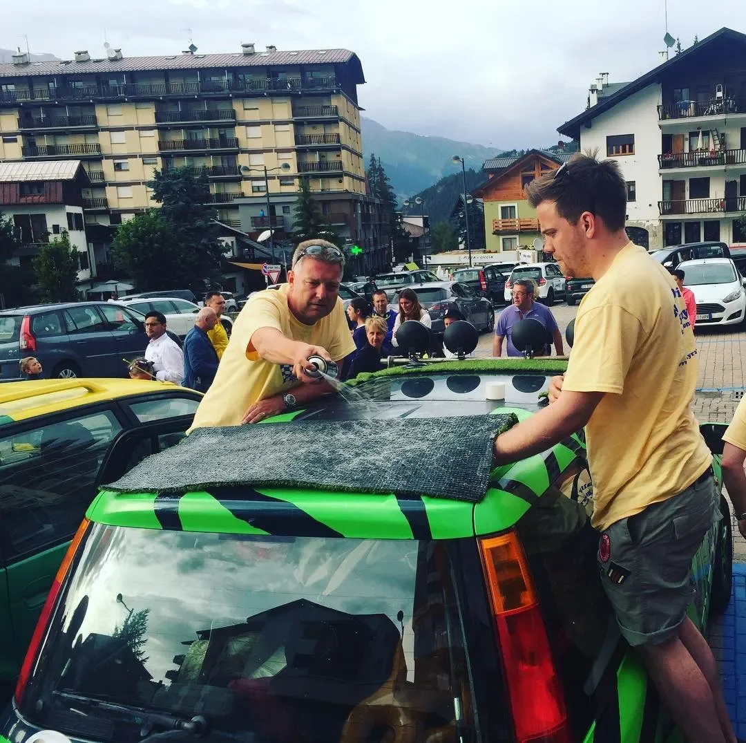 Two men in yellow shirts wash a small green car outdoors, surrounded by people and cars in a parking lot with buildings and mountains in the background.