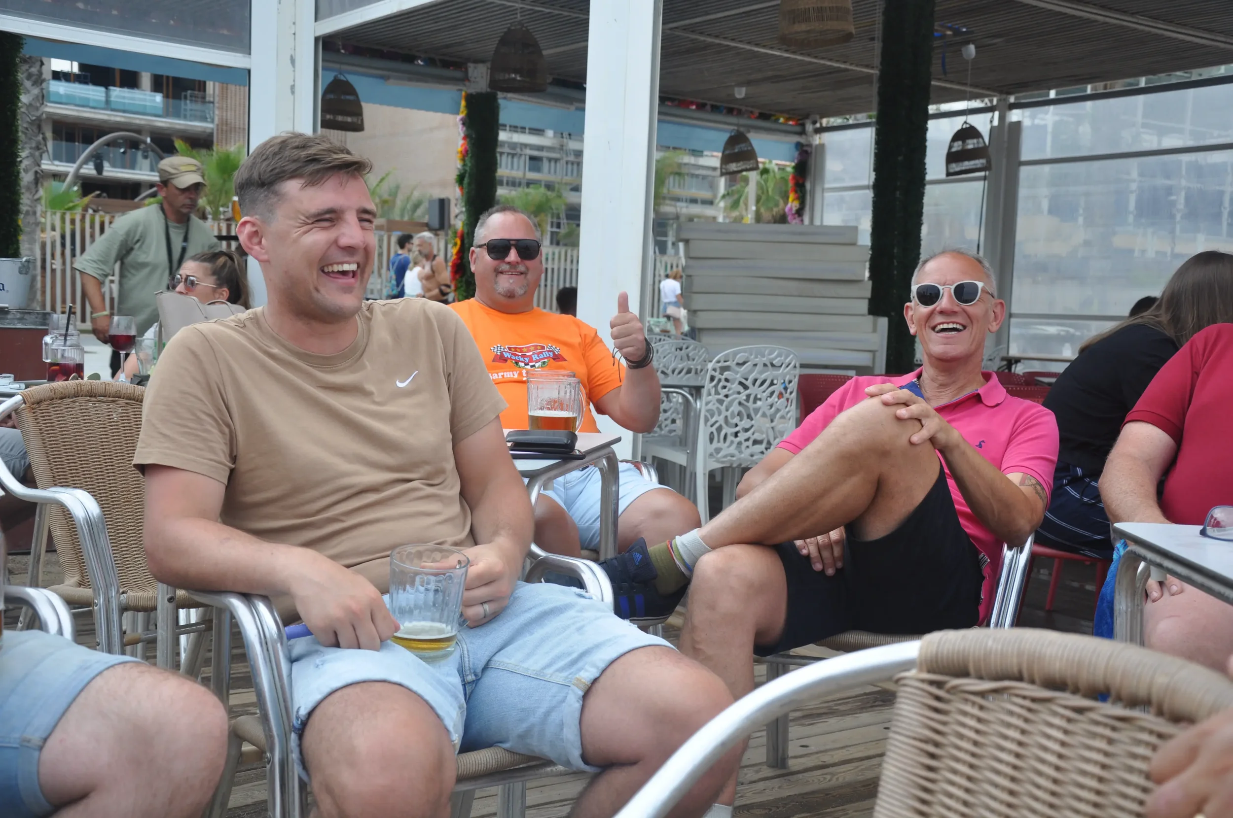 Group of people sitting and laughing at a social gathering with drinks, outdoors on a patio with modern decor.