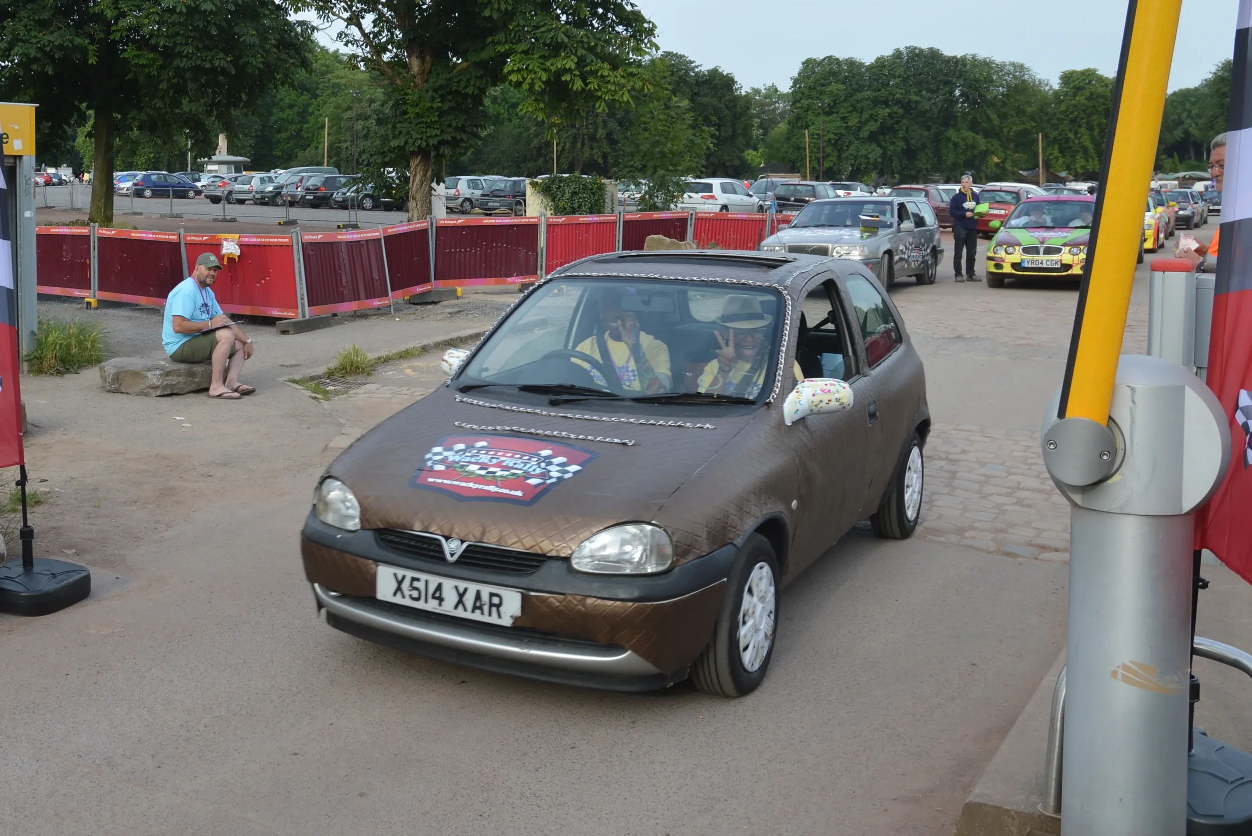 A small brown car with a checkered racing sticker on the hood and a custom license plateX514 XAR, driving towards a parking area on a street with trees in the background. Two people inside the car are smiling and making peace signs. Outside, a man is