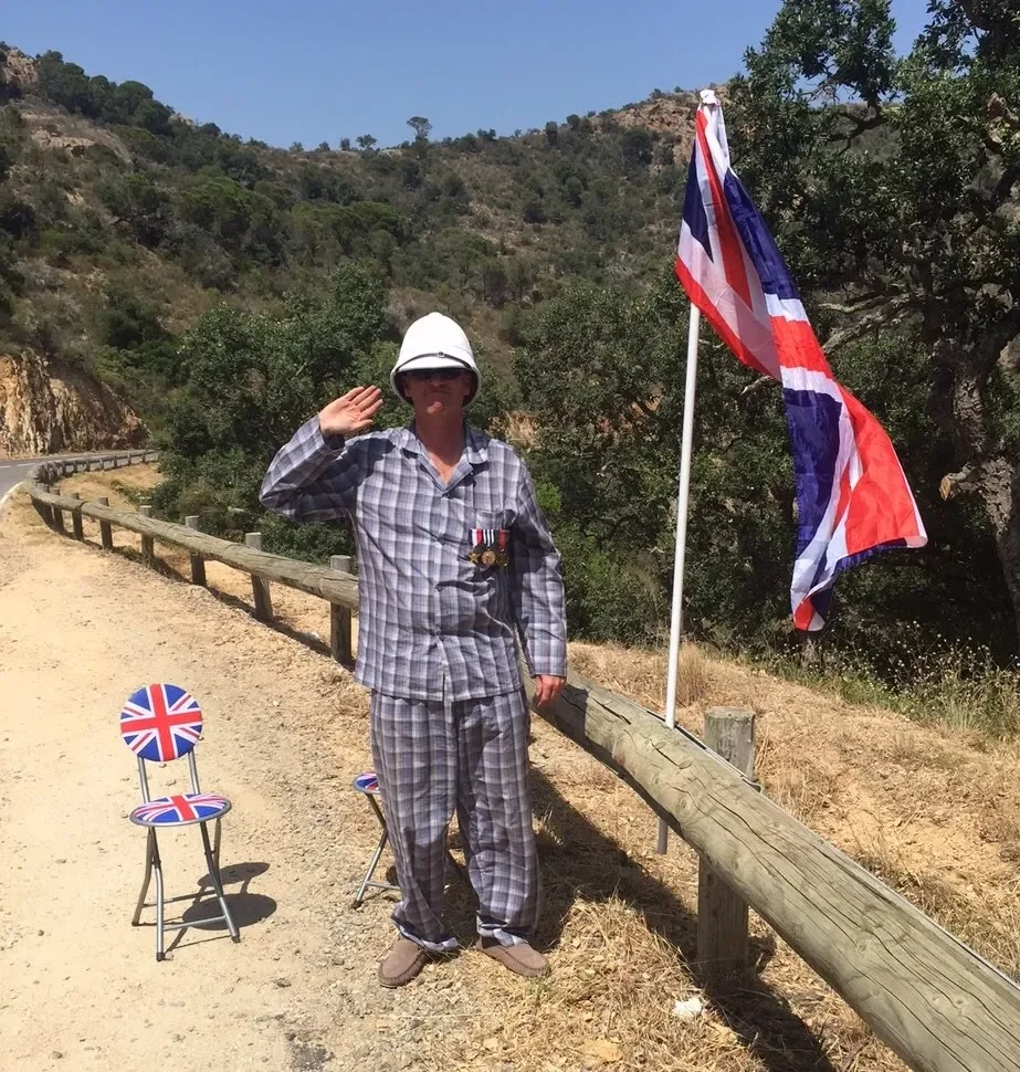 Person in plaid pajamas, wearing a white hard hat and medals, saluting next to a Union Jack flag and two small chairs with British flags on a dirt road surrounded by trees and hills.