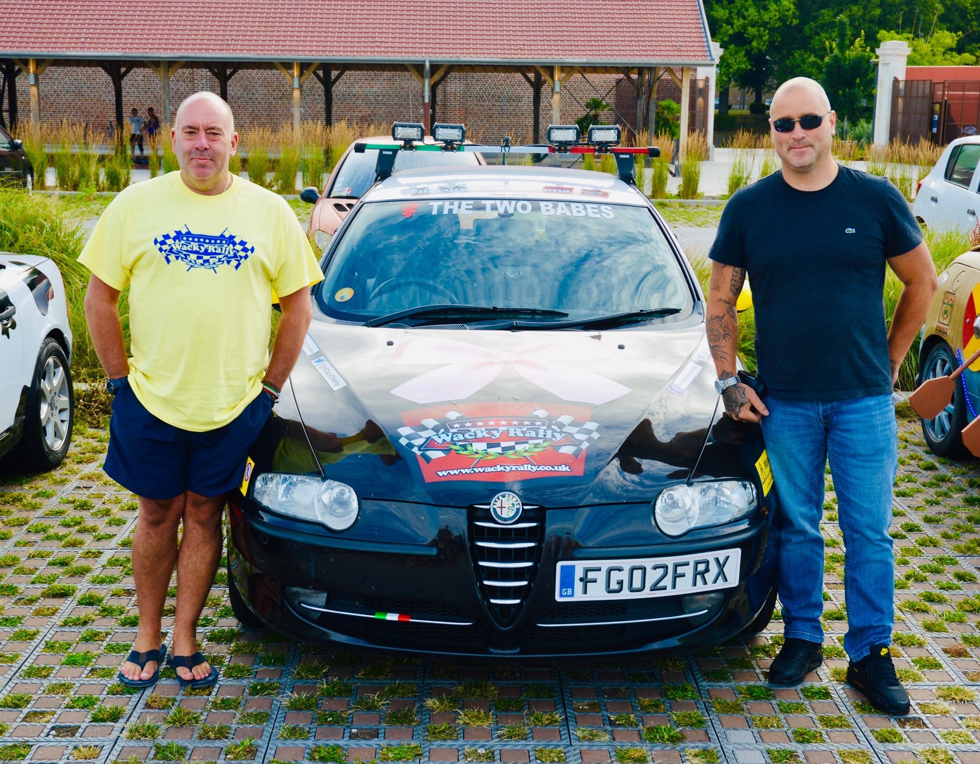 Two men standing on either side of a black Alfa Romeo car at a rally event. The car has a banner on the windshield reading 'THE TWO BABES' and a sticker on the hood for 'Wacky Rally' with a checkered flag logo and website link. The man on the left is