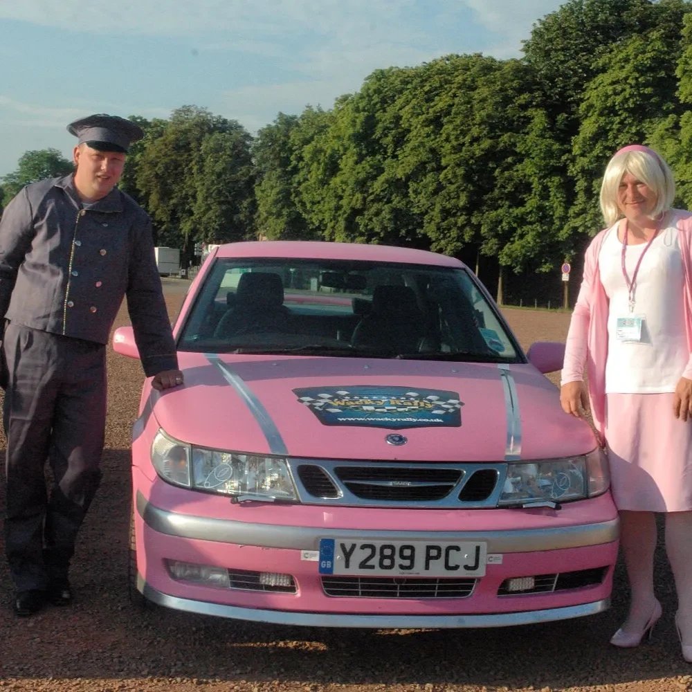 Two people dressed in costumes standing next to a pink car with a racing badge on the hood, in a parking lot surrounded by trees.