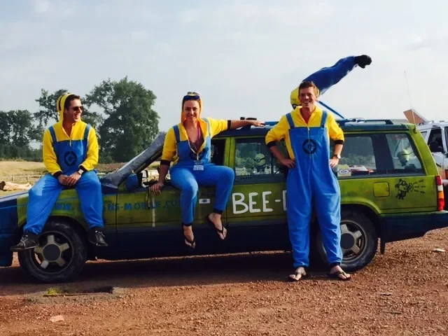 Three women wearing yellow and blue outfits with bee logos, sitting and standing on a green vehicle with 'BEE-' written on the side, outdoors with trees in the background.