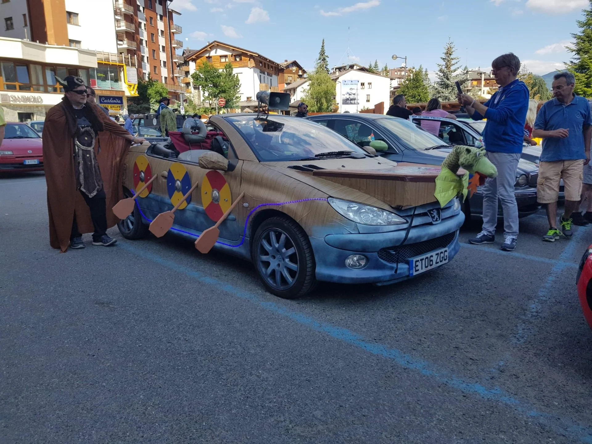A decorated car with a Viking warrior, shields, and swords, parked in a crowded outdoor event with people taking photos and a backdrop of buildings and trees.