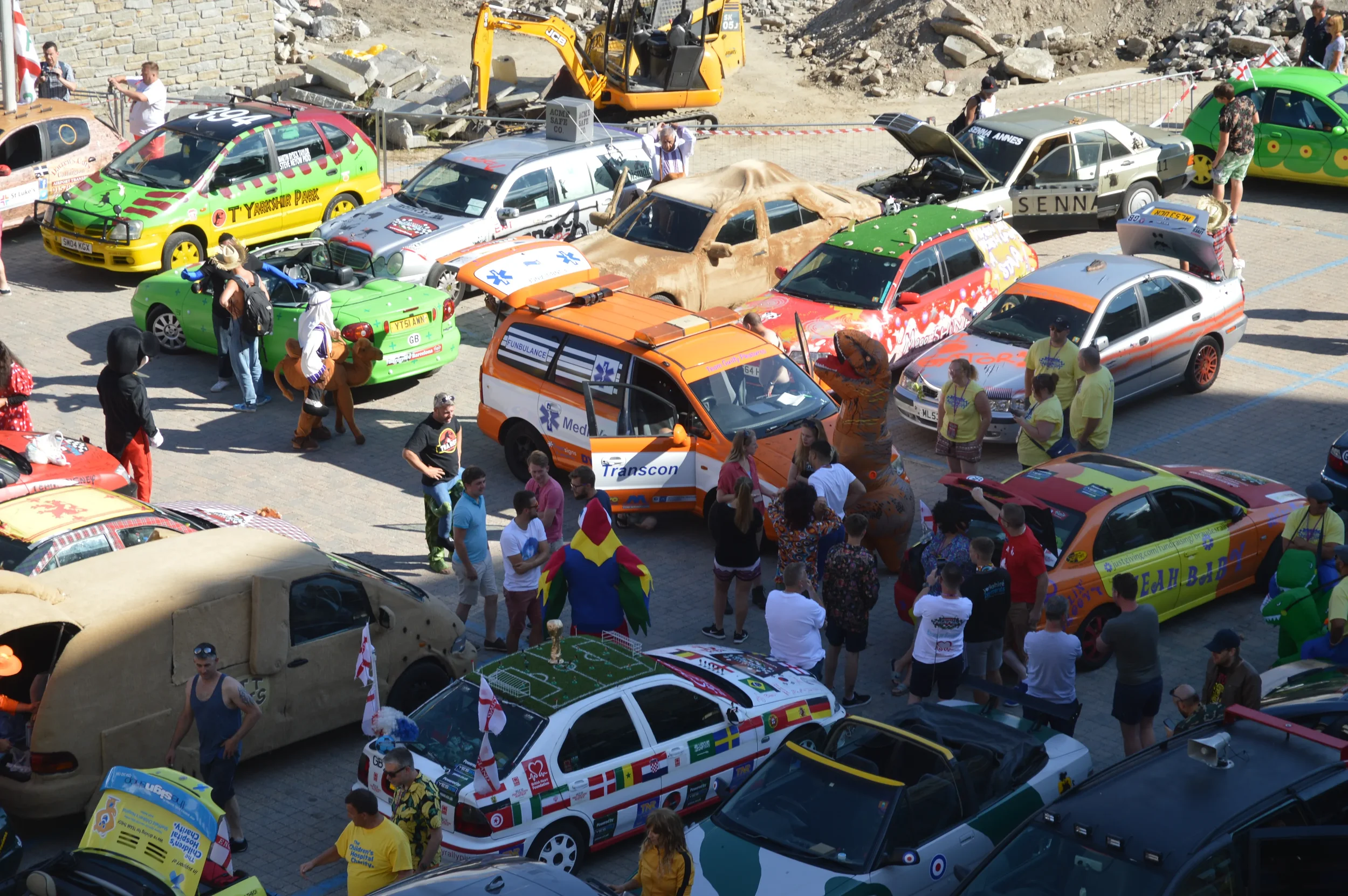 A car show with numerous decorated cars, people walking around, and some in costumes. Vehicles include race cars, themed cars, and a large inflatable dinosaur.