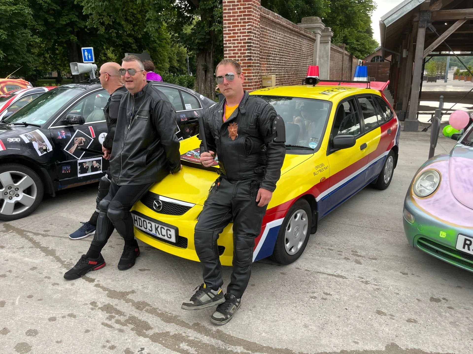 Two men in black leather racing suits and sunglasses leaning against a yellow police car with red and blue markings, parked on a concrete surface at an outdoor event.