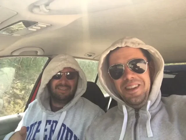Two men sitting in a car wearing hoodies and sunglasses, smiling at the camera.