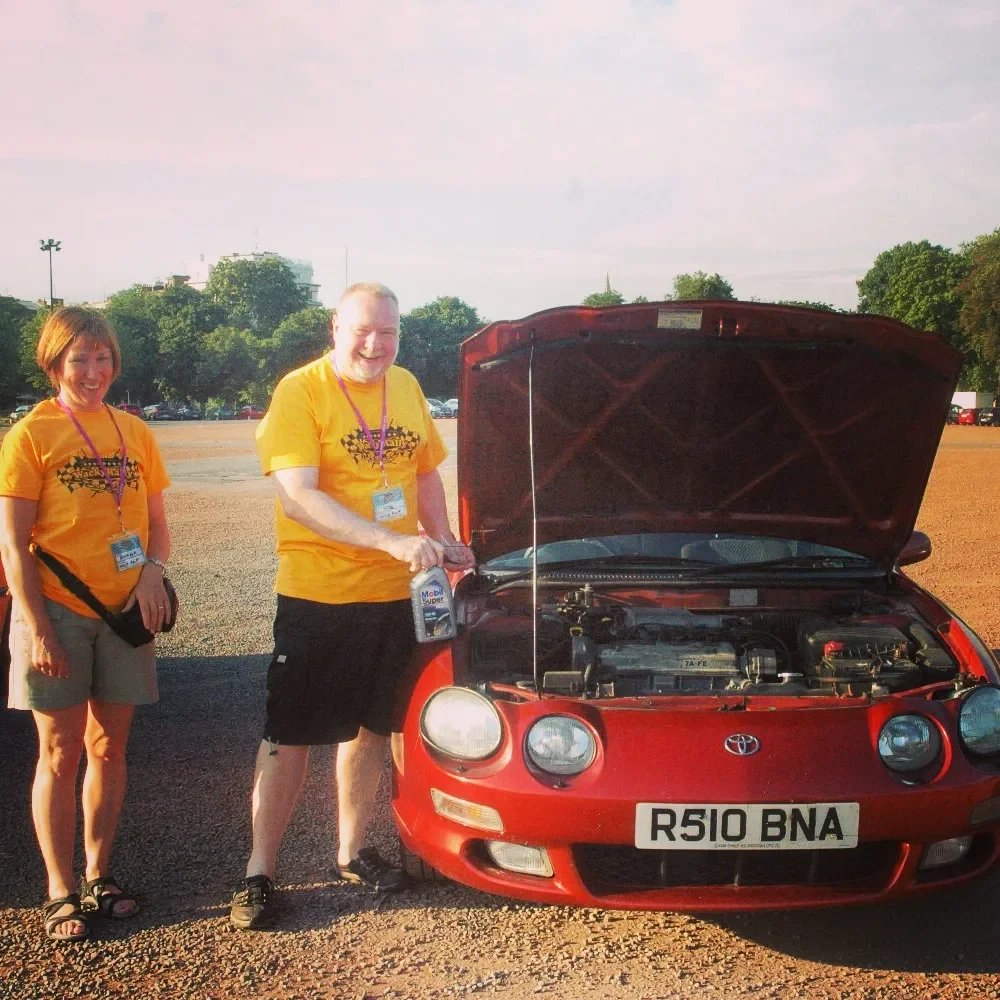 Two people in yellow t-shirts standing next to a red sports car with the hood open, in a parking lot.