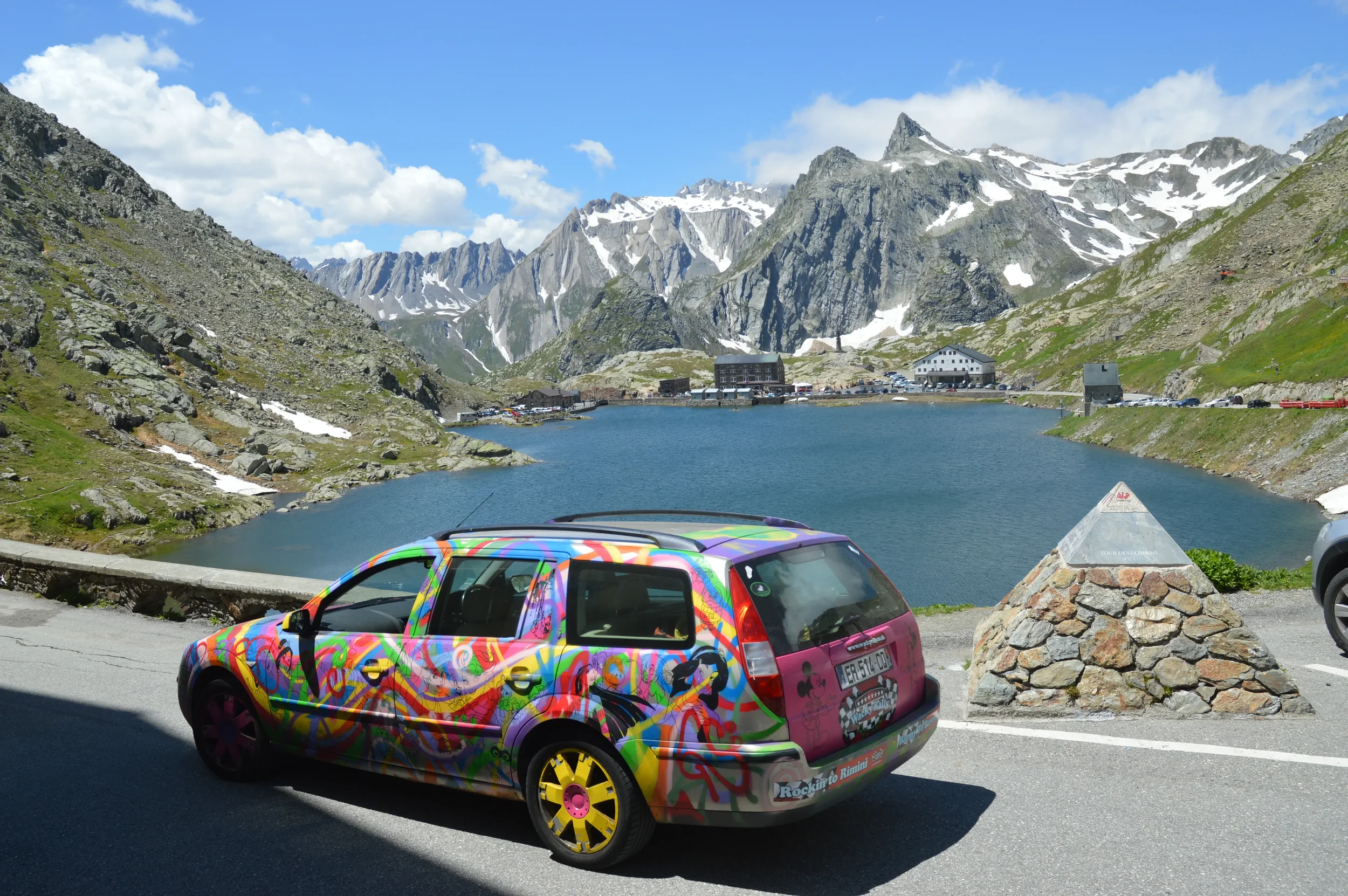 Colorfully painted car parked by a mountain lake with snow-capped peaks in the background.