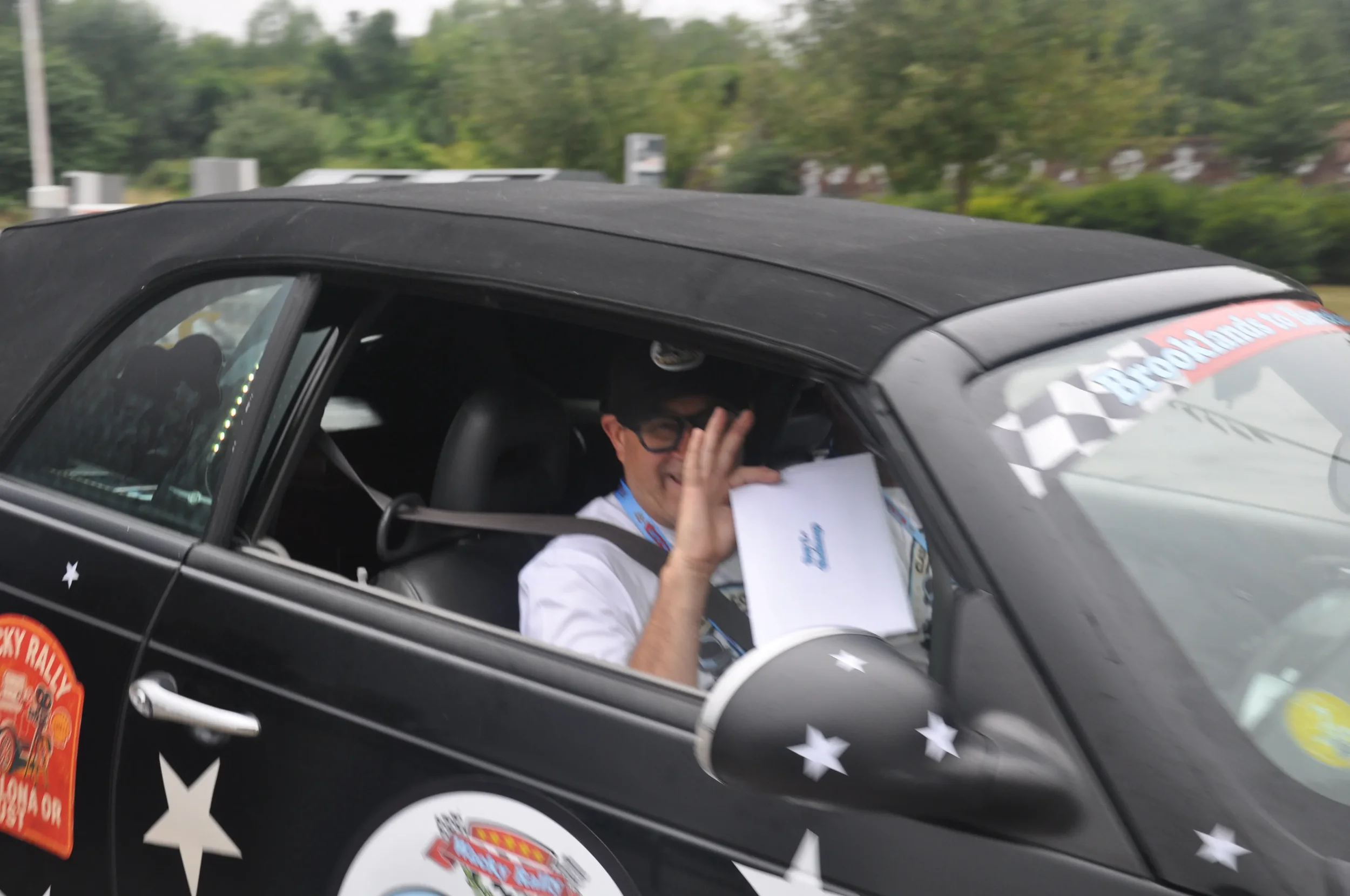 Person smiling and waving from inside a black rally car with stickers, holding a certificate or document. The car is outdoors with trees in the background.