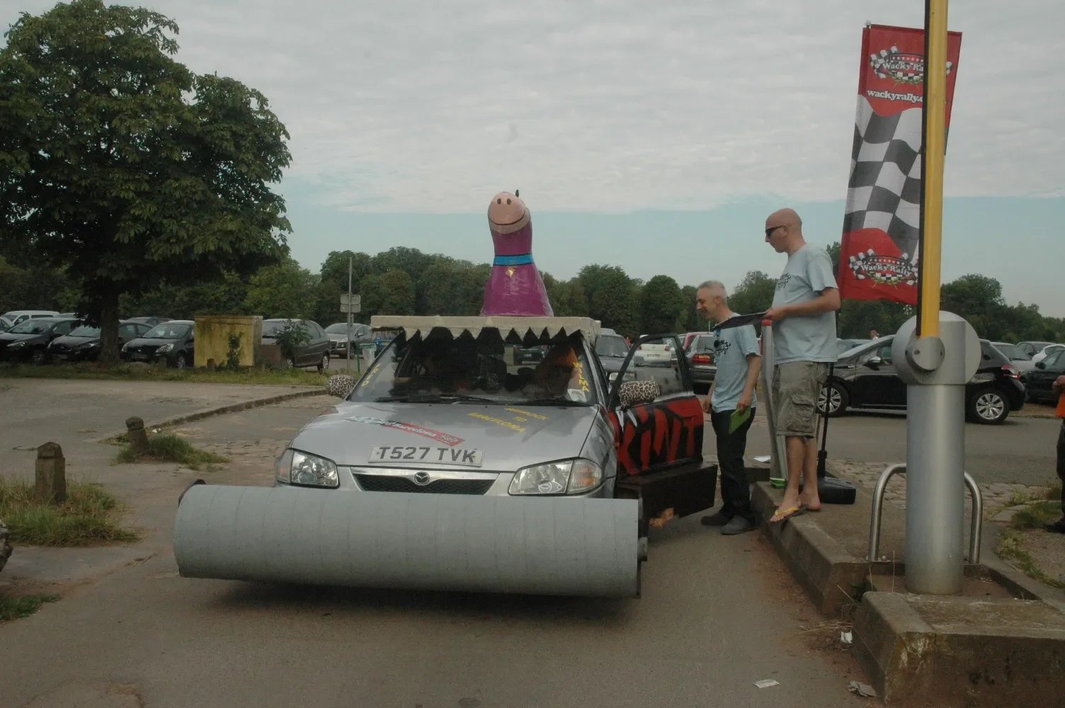 A car decorated with a large Wacky Races character at a parade or event, with people standing nearby and a parking lot in the background.
