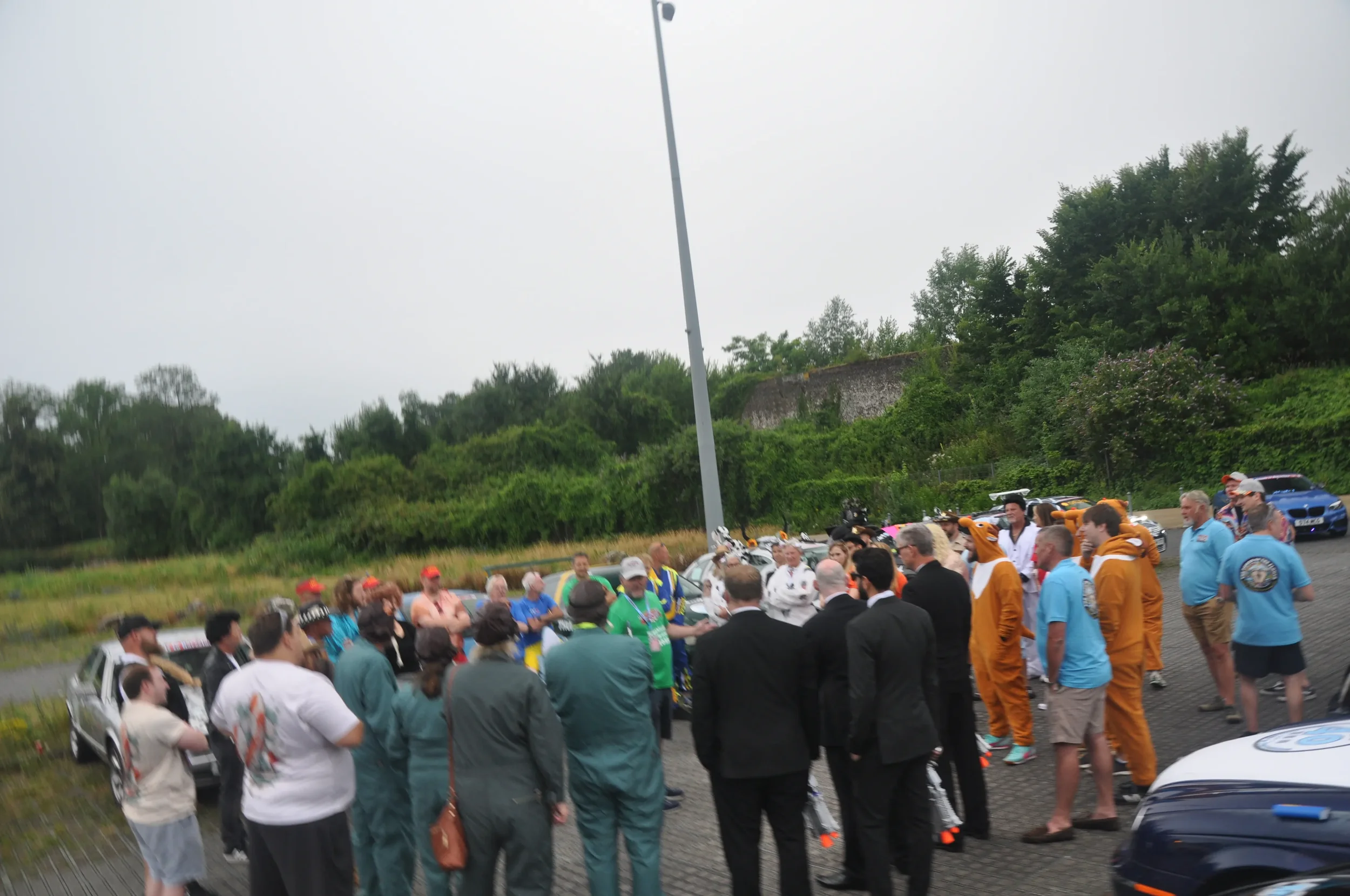 Group of people gathered outdoors, some in race car driver suits, others in casual or formal attire, near parked cars on a cloudy day, with trees and greenery in the background.
