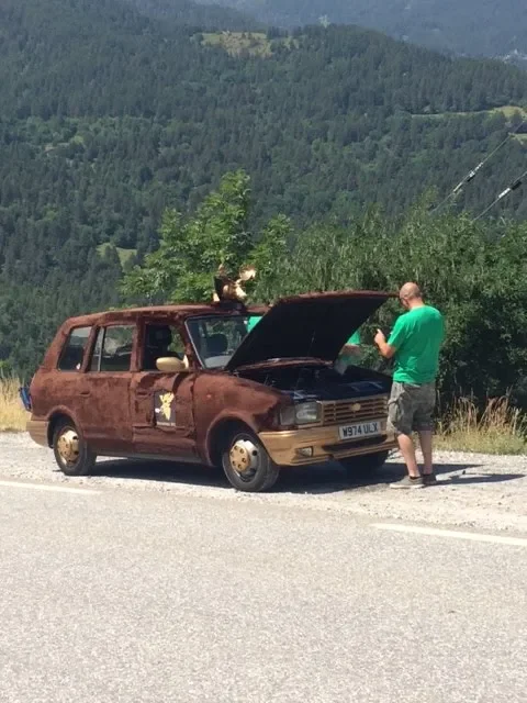 A rusty, old station wagon with its hood open, parked on the side of a rural road. A man in a green t-shirt and shorts is standing next to the car, looking at his phone. A small dog is sitting on top of the car, near the open hood, with a scenic moun