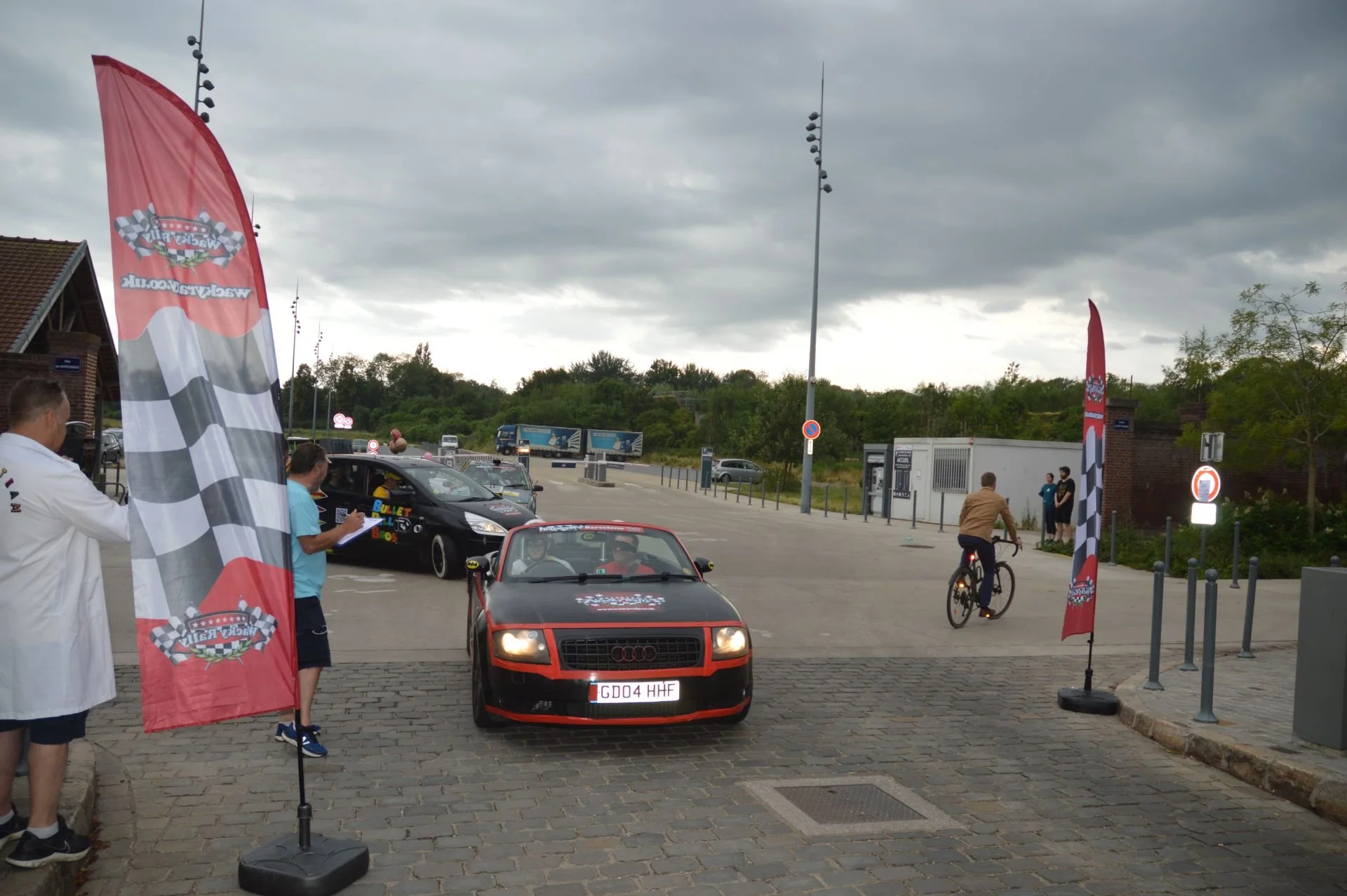 A racing event setup with red and black Audi car in the foreground, a black car behind it, and two flags with racing checkered patterns and logos on either side. People are present, one taking notes and another riding a bicycle, with cloudy skies abo