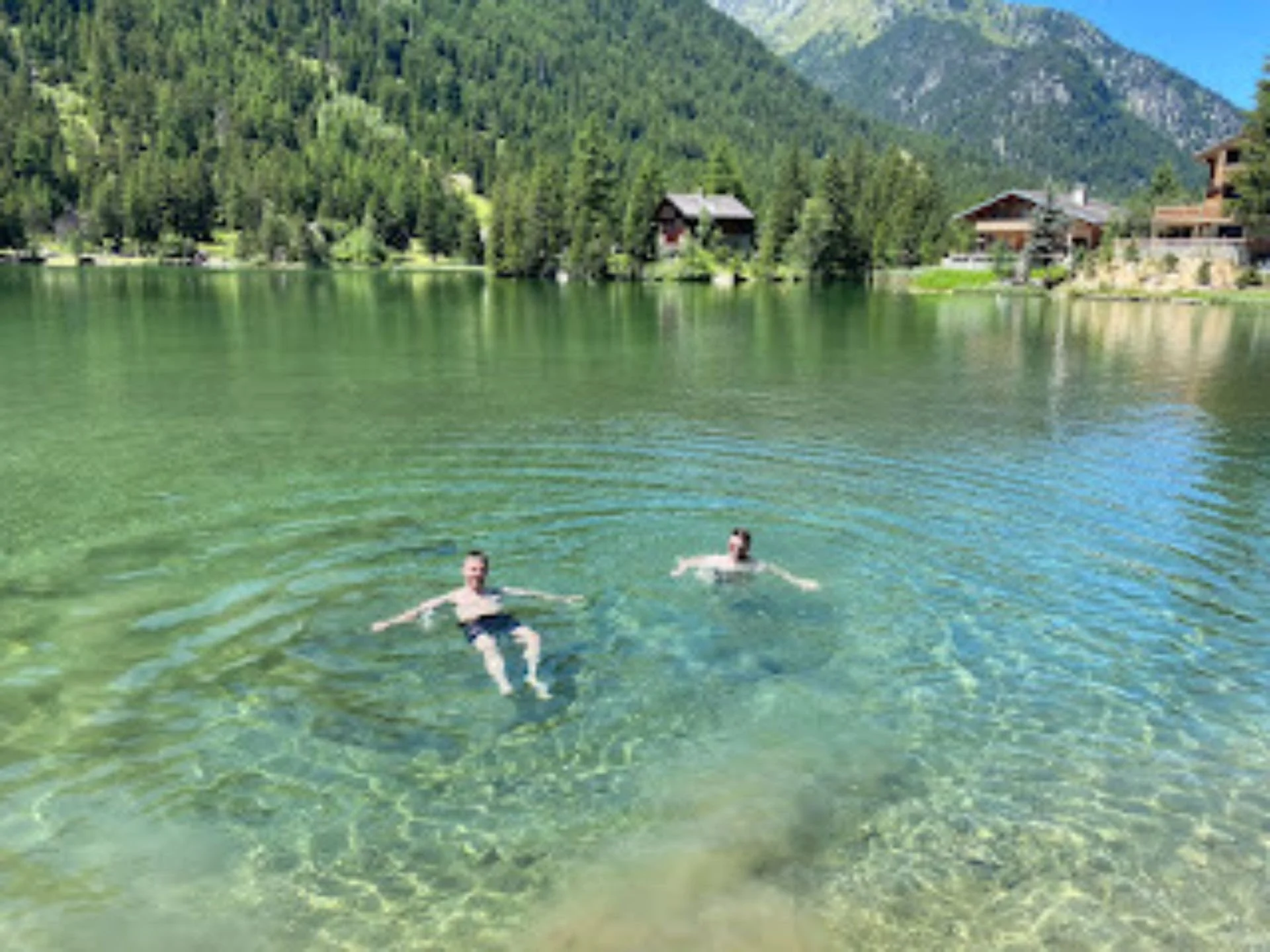 Two people swimming in a clear mountain lake with houses and green trees in the background.