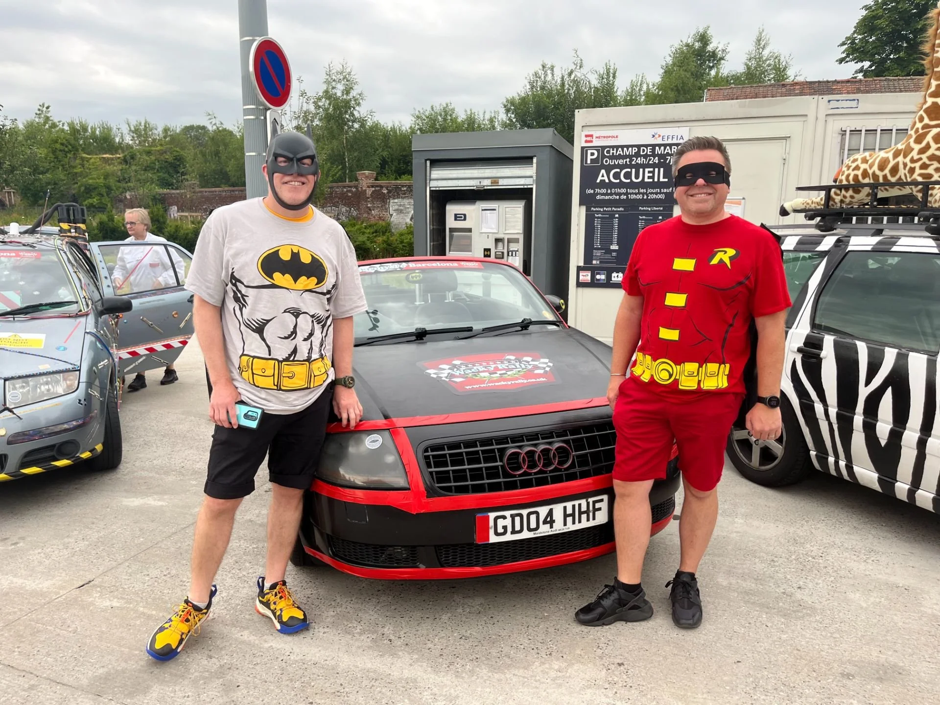 Two men wearing superhero masks dressed as Batman and Robin standing next to a black and red Audi car with custom license plate, in a parking lot with other decorated cars in the background.
