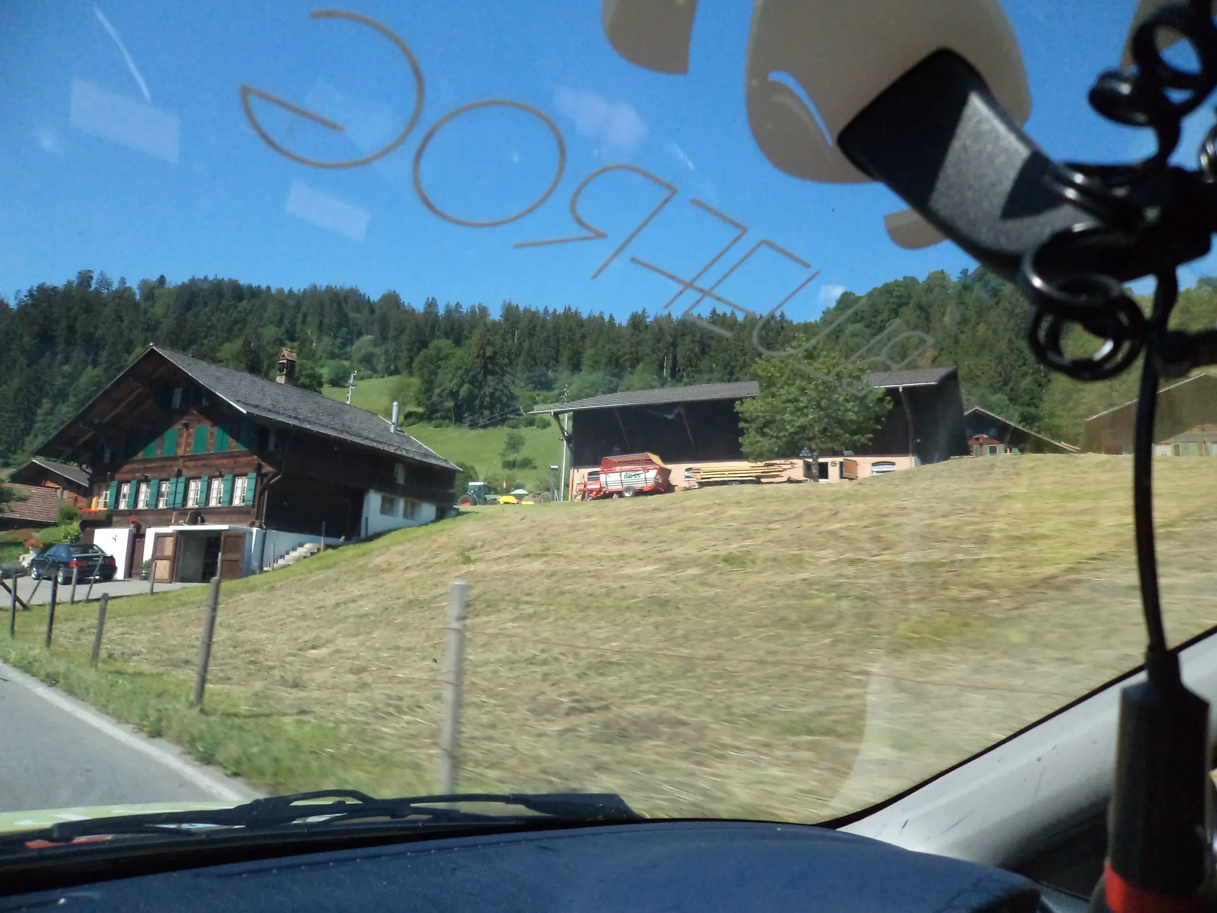 View of houses and a hillside seen through a vehicle windshield, with some reflections and objects hanging inside the car.