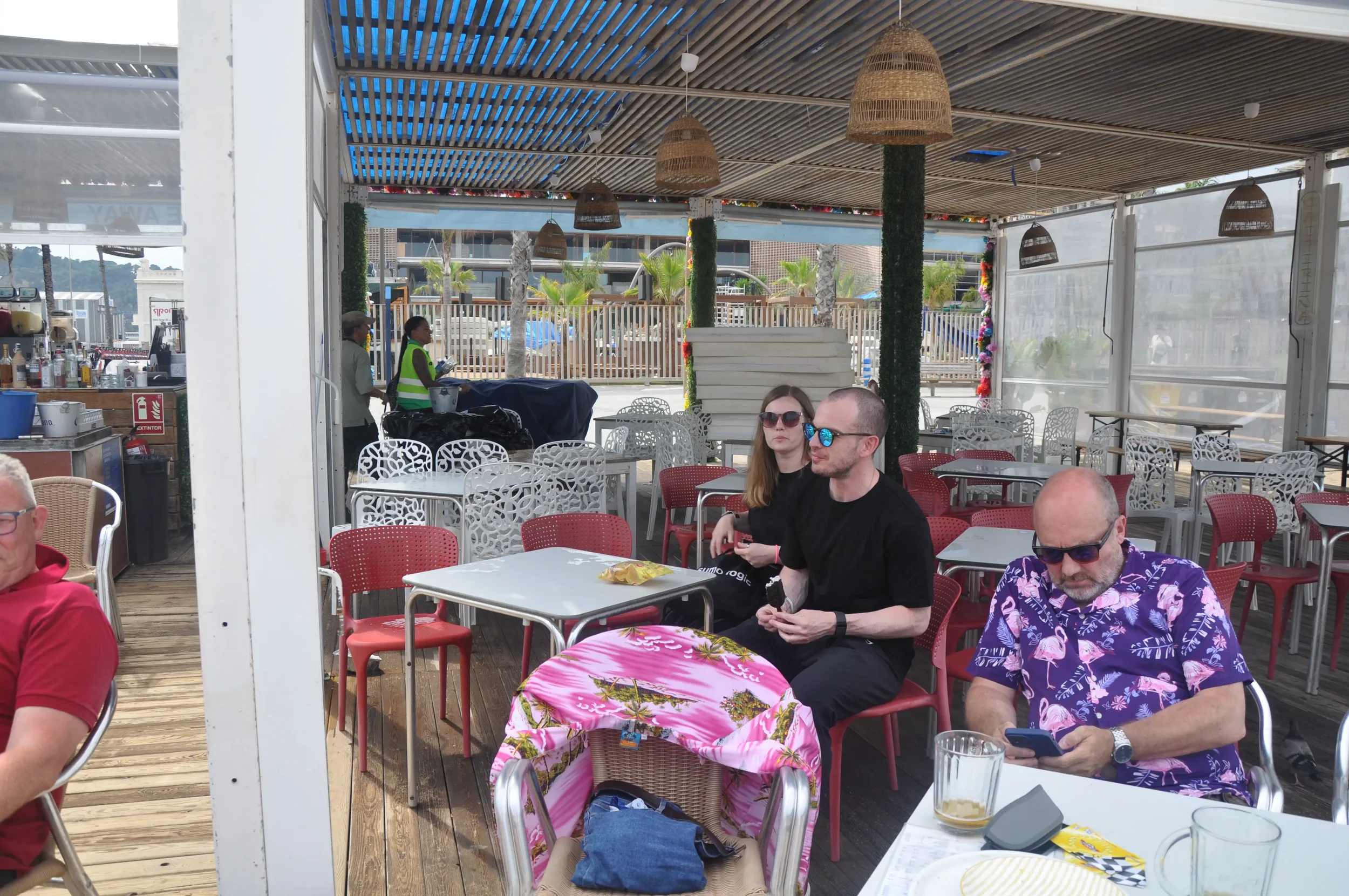 People sitting at outdoor restaurant or cafe with tables, chairs, and sunglasses, some looking at phones, under a shaded canopy.