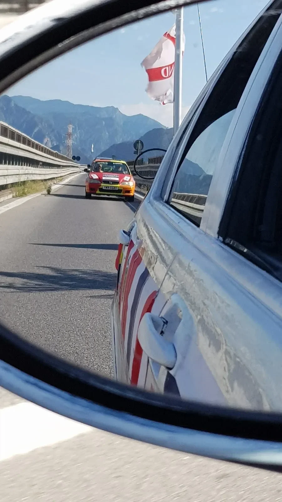 A race car with a yellow front on a mountain highway, viewed through a side mirror of a car, with a red, white, and yellow race car ahead, and mountains in the background.