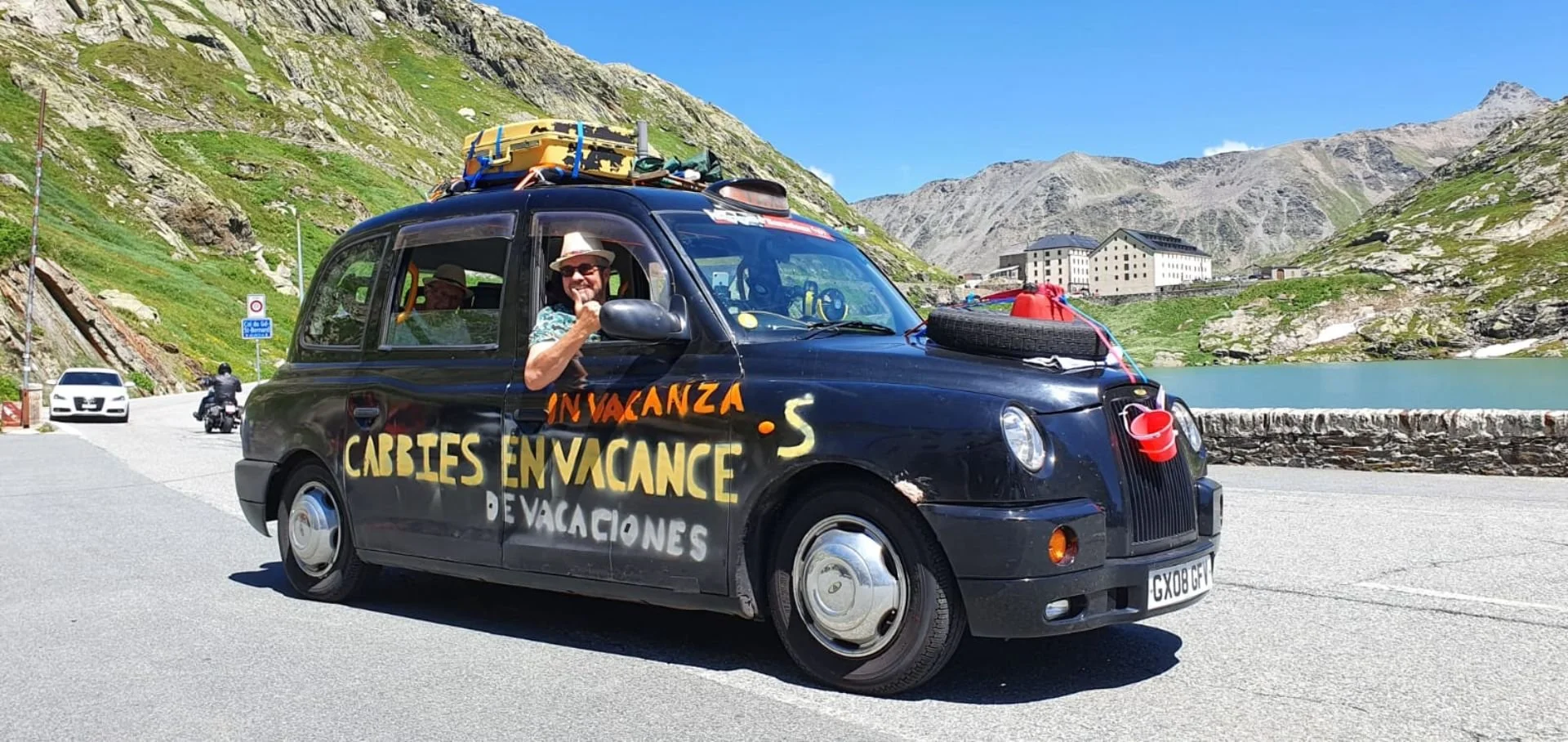 A black car parked near a mountain lake with a man smiling and giving a thumbs-up from the driver's side window. The car is decorated with colorful writing in Spanish promoting a vacation in Cabbies. It has luggage on the roof and various camping sup