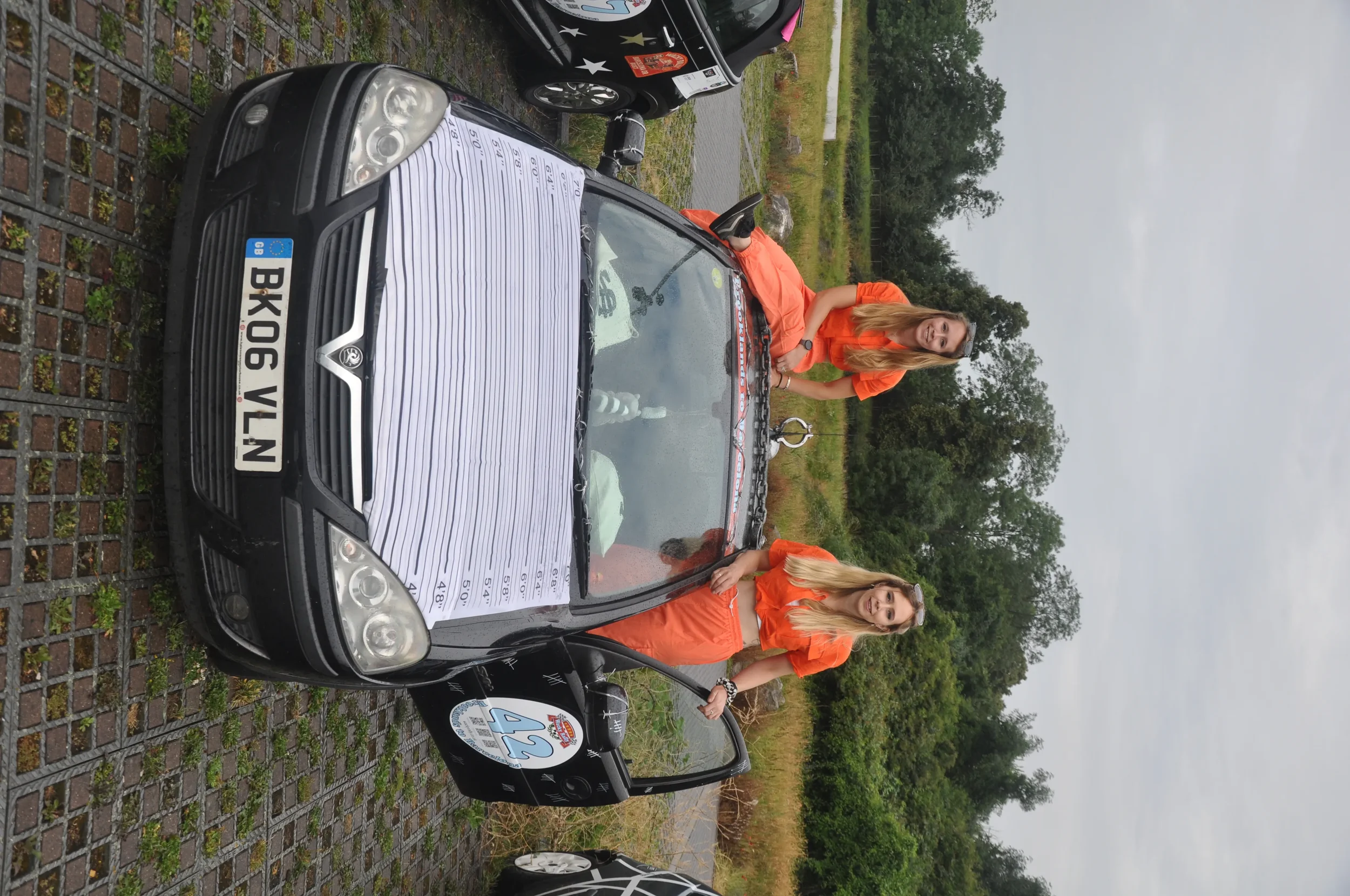 Two young women in orange shirts standing next to a black car with a covering on the hood. The car is parked on a gravel path near grassy and wooded areas, under an overcast sky.