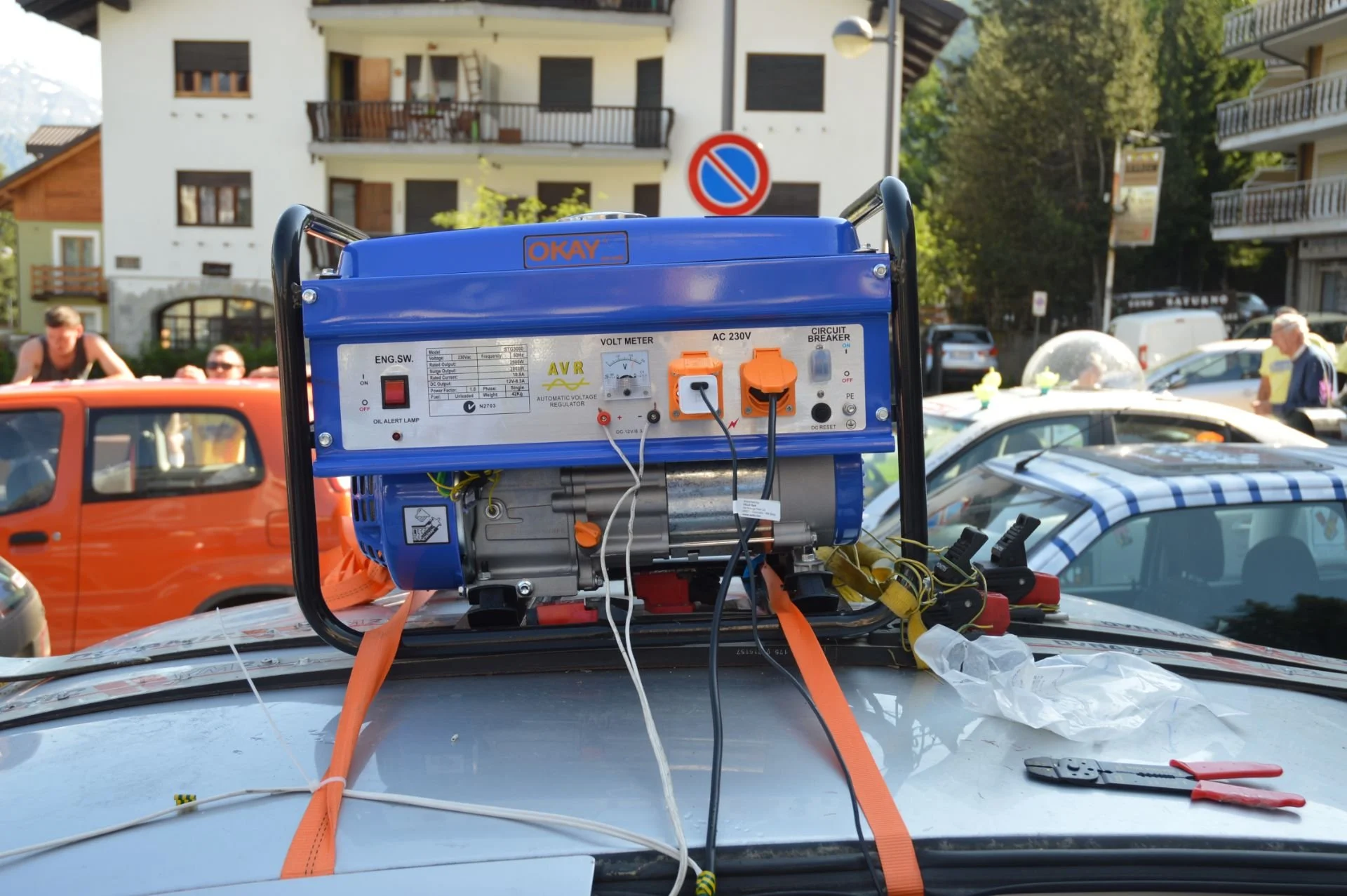 A portable generator placed on a car roof with various cables connected, set in a parking lot with several cars and buildings in the background during daytime.