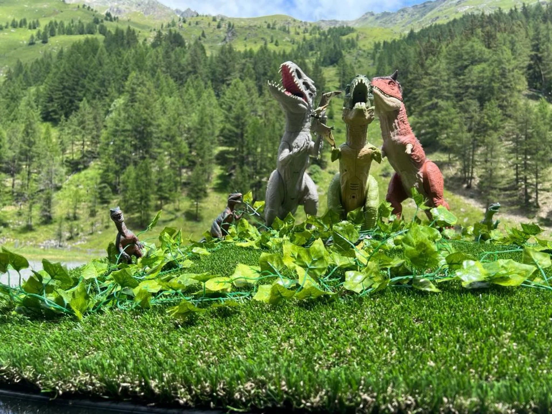 Dinosaur figurines and toy plants on a grassy patch with a forest and mountain background.