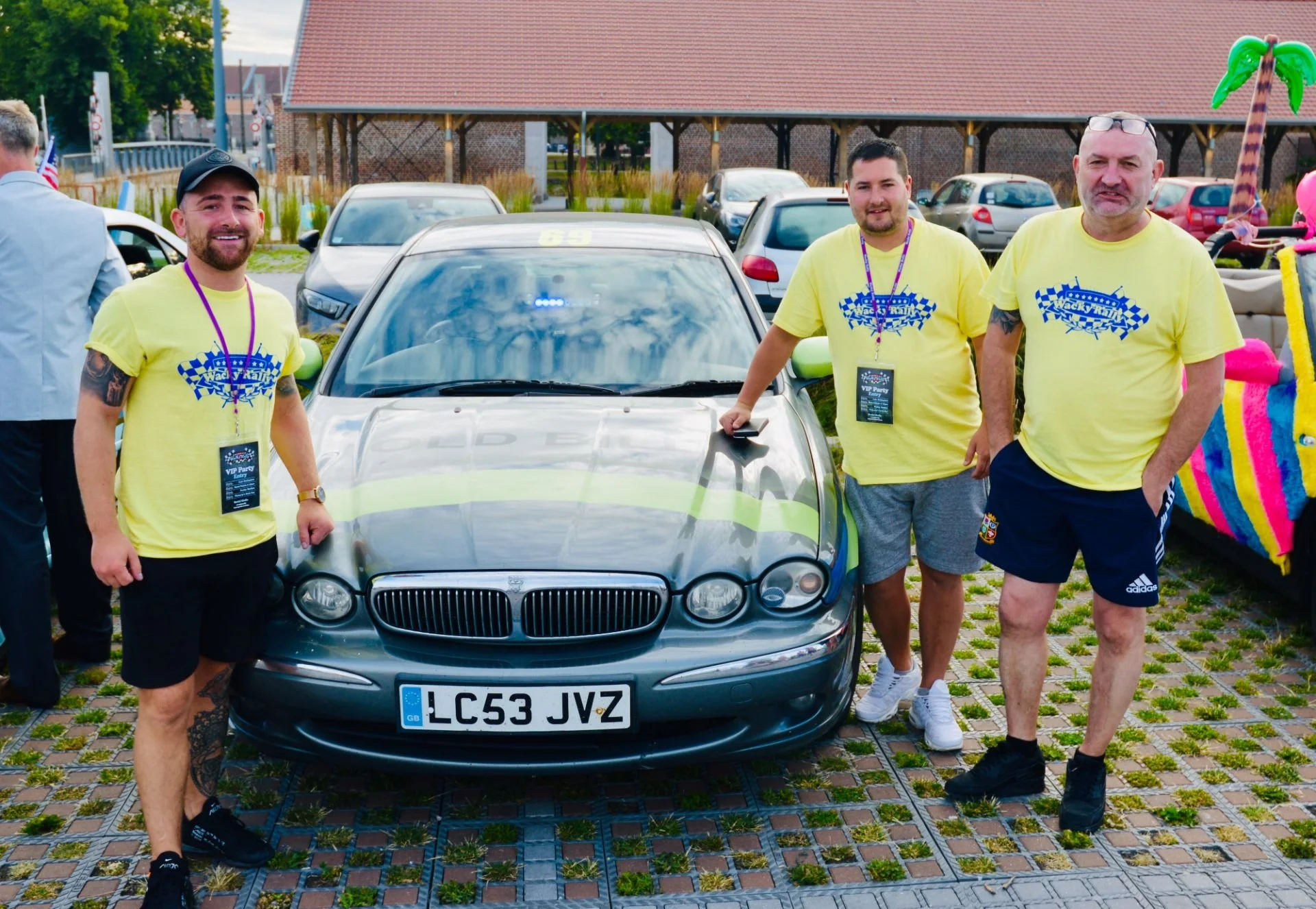 Three men standing around a black car at an outdoor event, wearing yellow t-shirts with a racing logo, with a colorful inflatable decoration in the background.