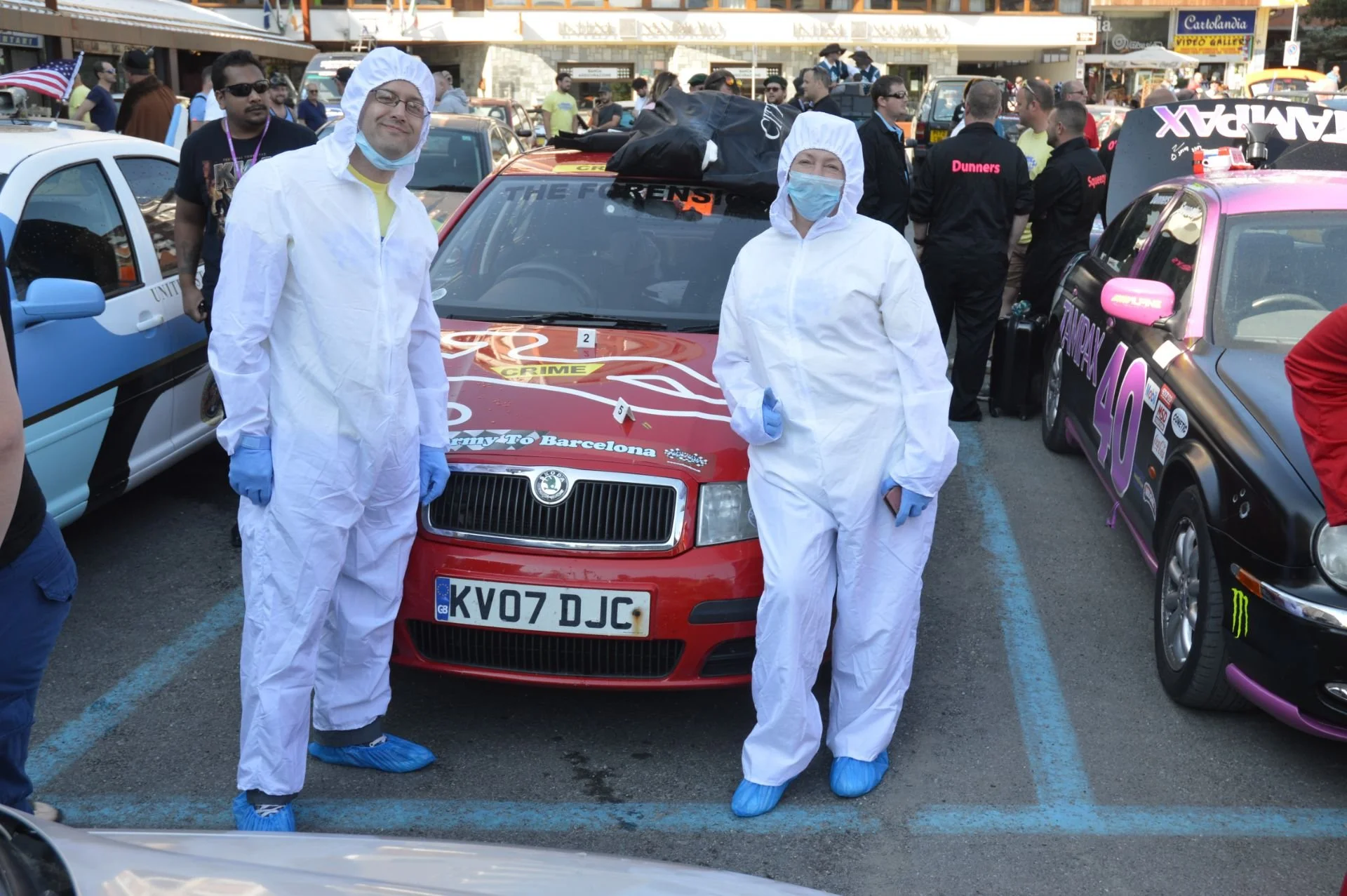 Two people dressed in white protective suits, face masks, and gloves standing in front of a red rally car with the license plate 'KV07 DJC'; they are at a racing event with other cars and people in the background.