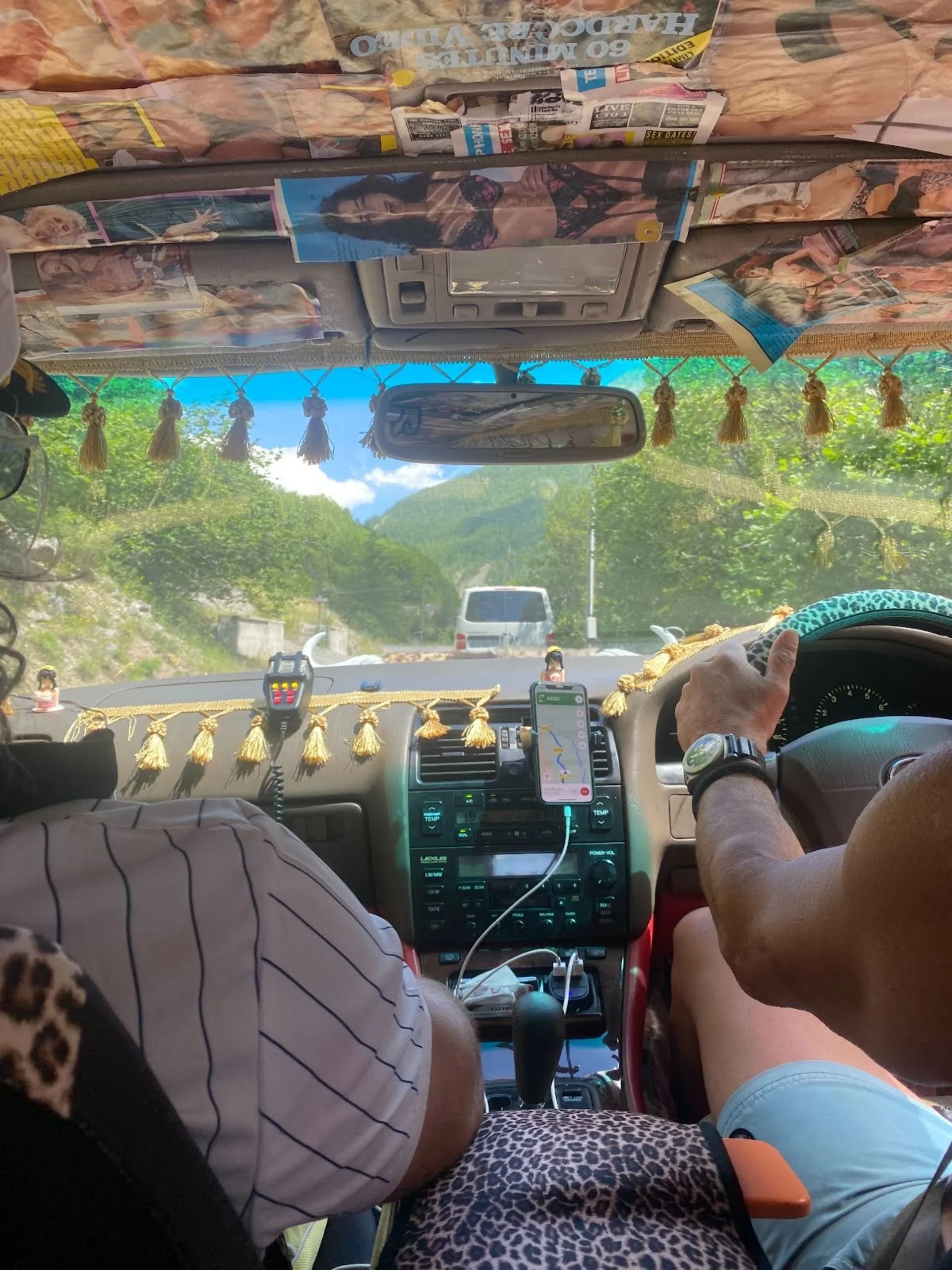The image shows the interior of a vehicle with two passengers. The dashboard is decorated with tassels, and there is a GPS and a walkie-talkie on the dashboard. The windshield reveals a scenic view of a green, hilly landscape with a road and a mounta
