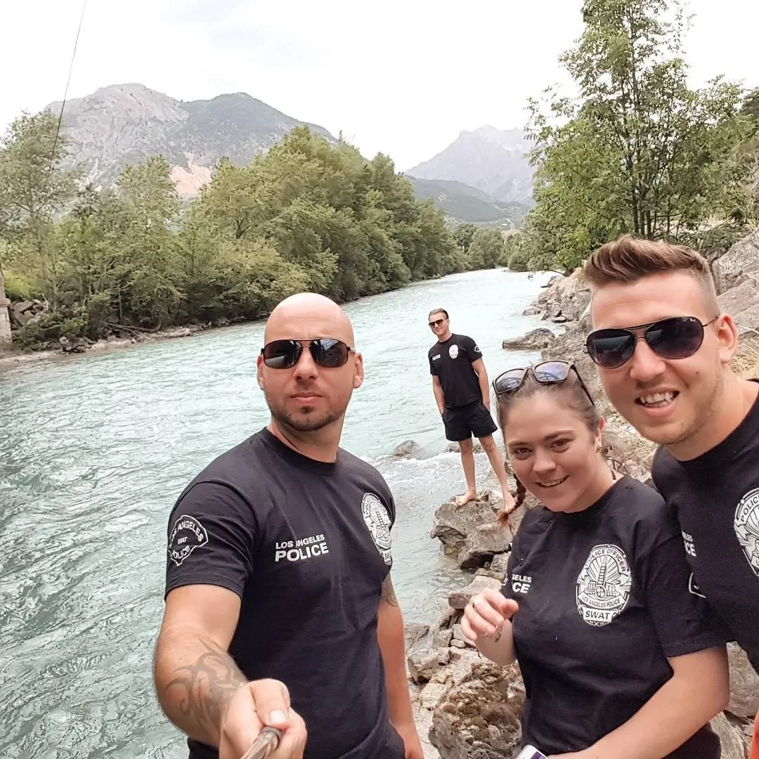 Four police officers in uniform taking a selfie on rocks beside a river with mountains and trees in the background.