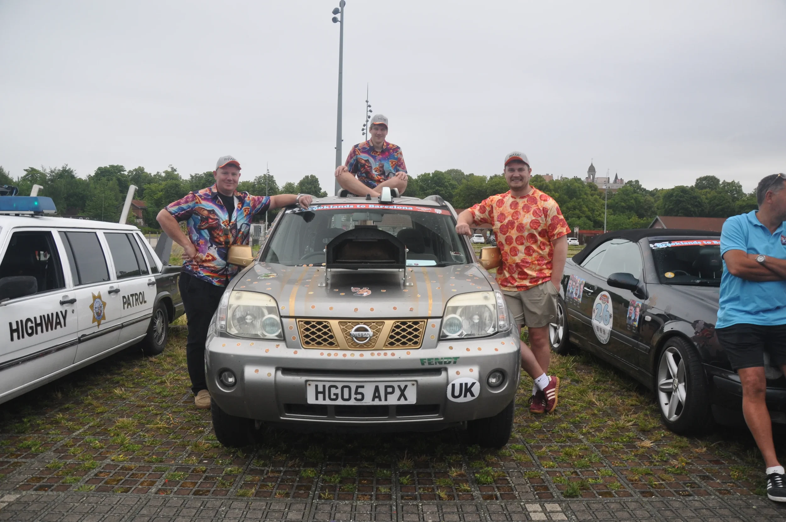 Three men standing and sitting around a decorated Nissan SUV at an outdoor event, with classic cars and a police patrol car nearby, overcast sky.