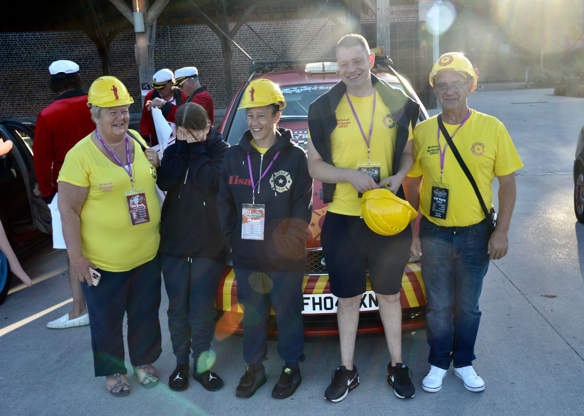 Group of people standing in front of a red car, wearing yellow safety helmets and yellow shirts, some with event badges, during a daytime outdoor event.