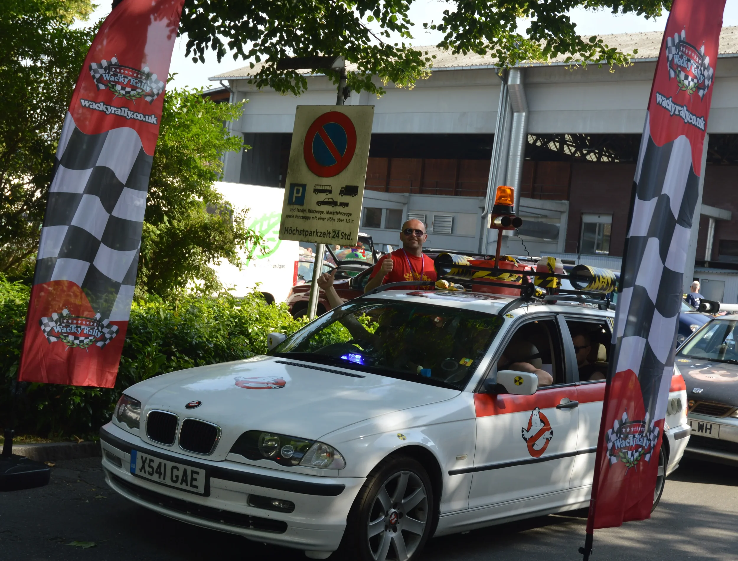 A white car decorated for a rally event, with Wacky Rally banners on both sides, and a man wearing sunglasses and a red shirt giving a thumbs-up while standing behind the car. The car has various equipment and a Ghostbusters logo sticker on the door.