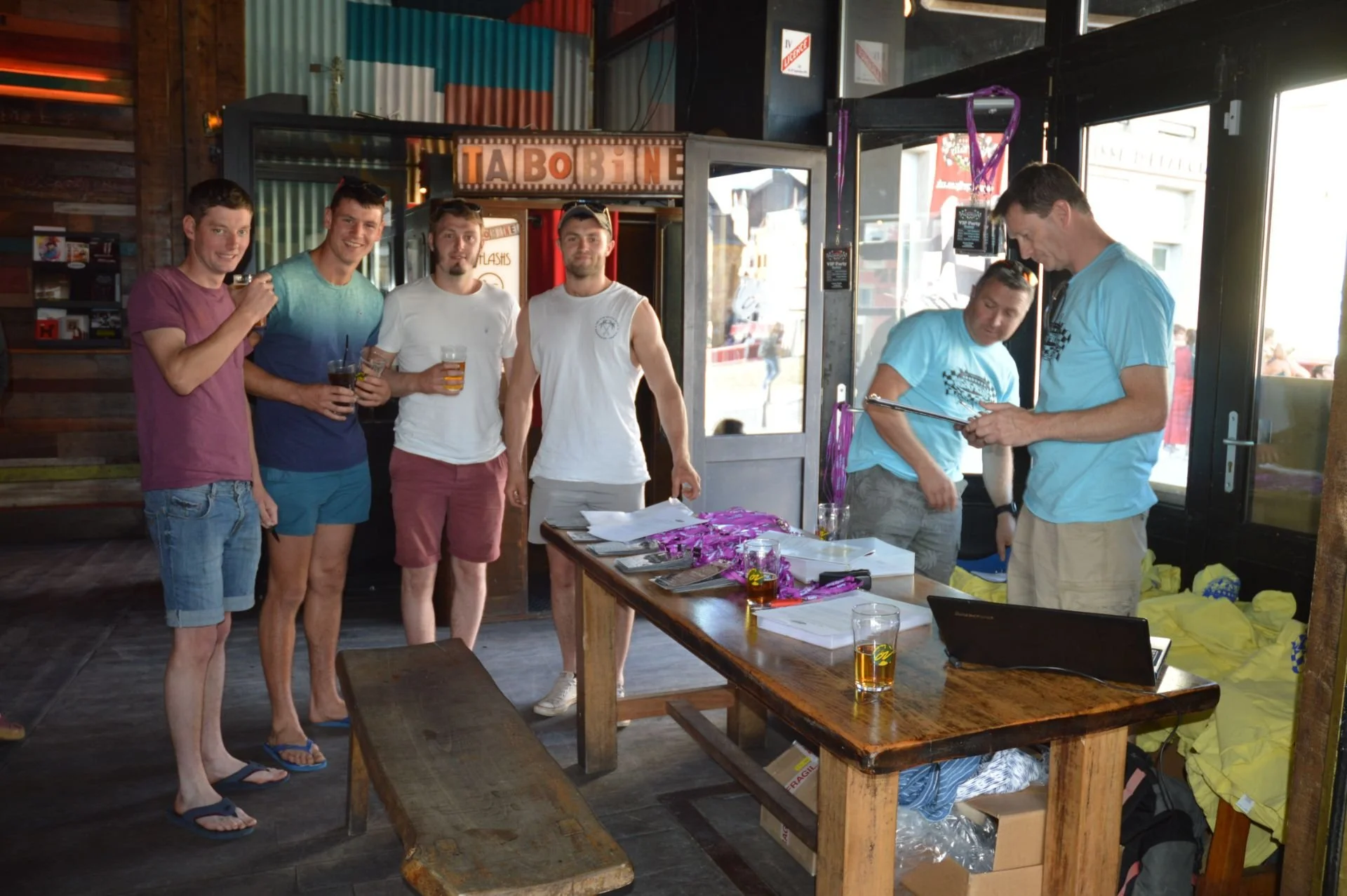 A group of five young men standing inside a bar, smiling, with some holding drinks, and two other people behind a table, looking at papers; the bar has a rustic style with colorful tin paneling and wooden furniture.