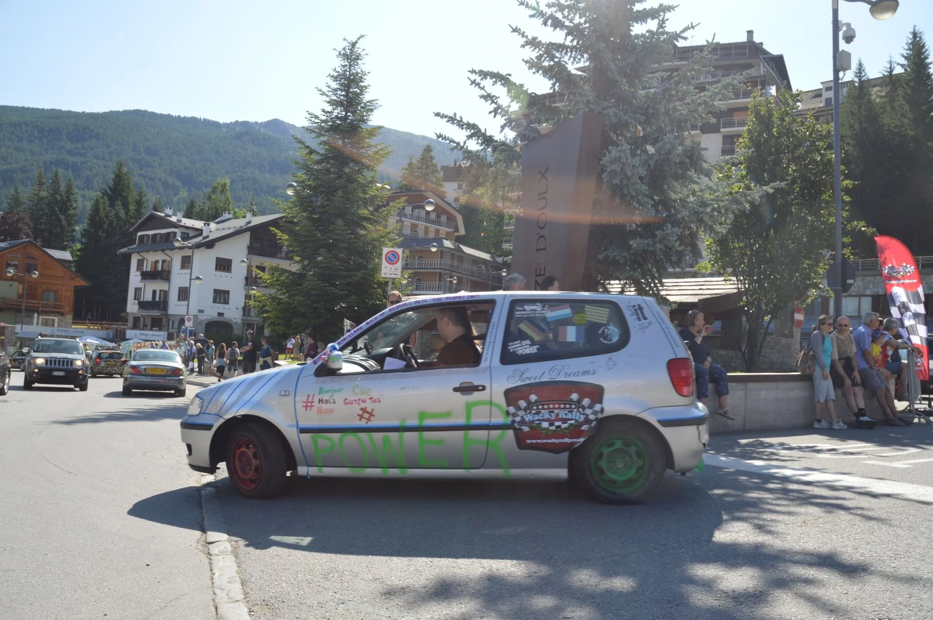 A decorated car with the word 'POWER' written on the side, driving through a street in a mountain town with people sitting and standing around, trees, and buildings in the background.