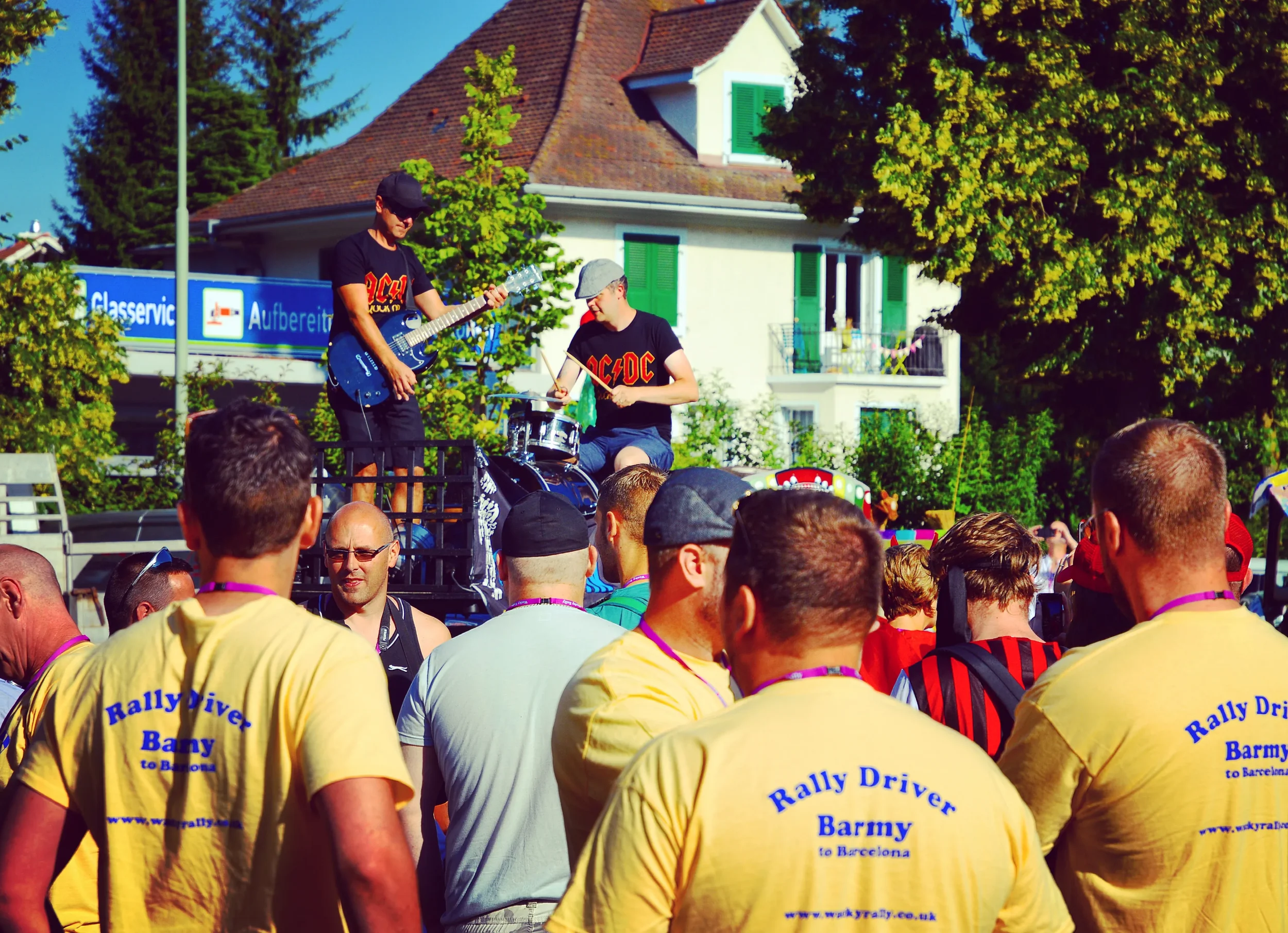 People attending a live outdoor concert with a band performing on stage, and some of the attendees are wearing yellow shirts with 'Rally Driver Barmy to Barcelona' written on the back.